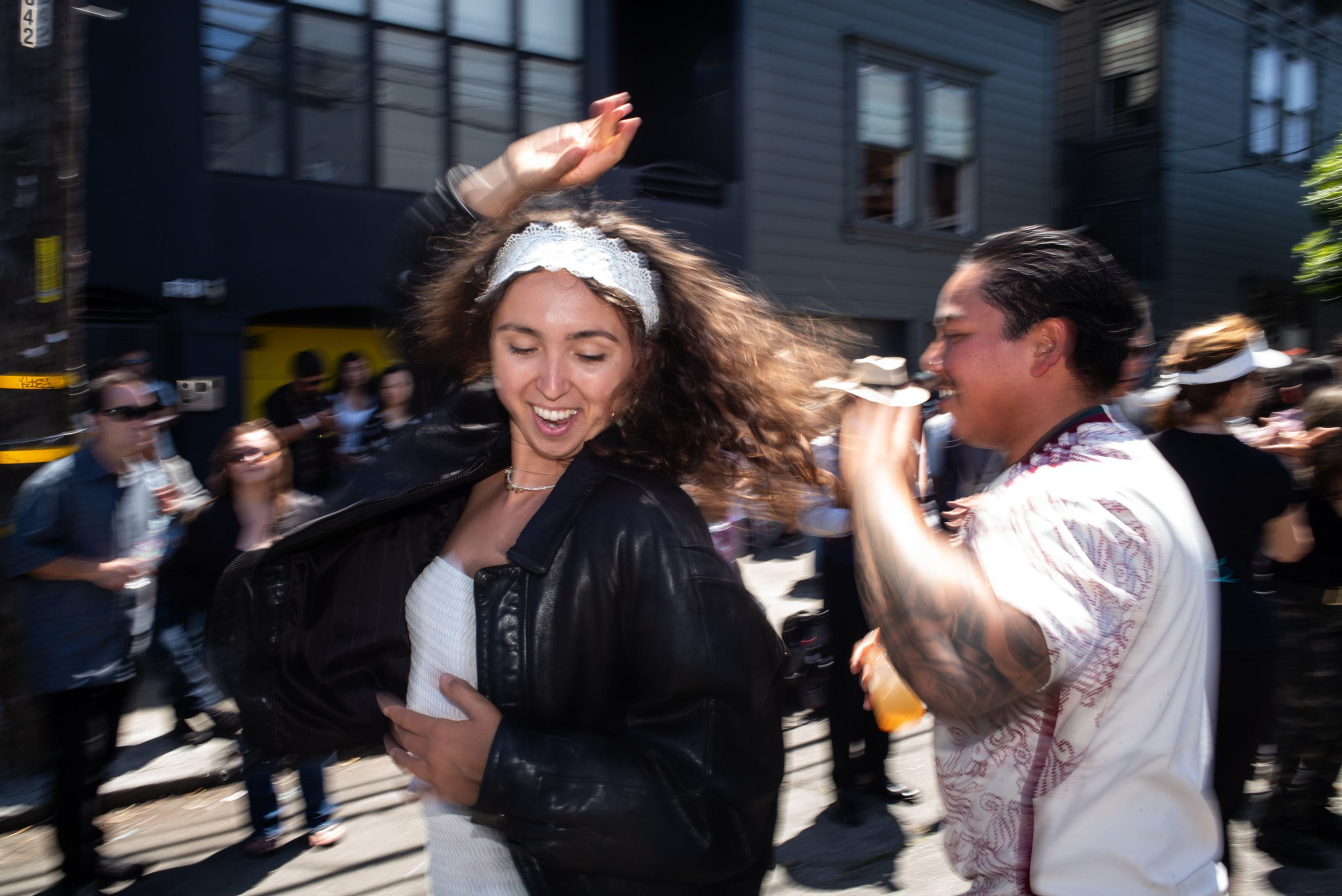 A woman with curly hair and a white headband dances joyfully while a man with tattoos and a drink watches her, surrounded by people outdoors.