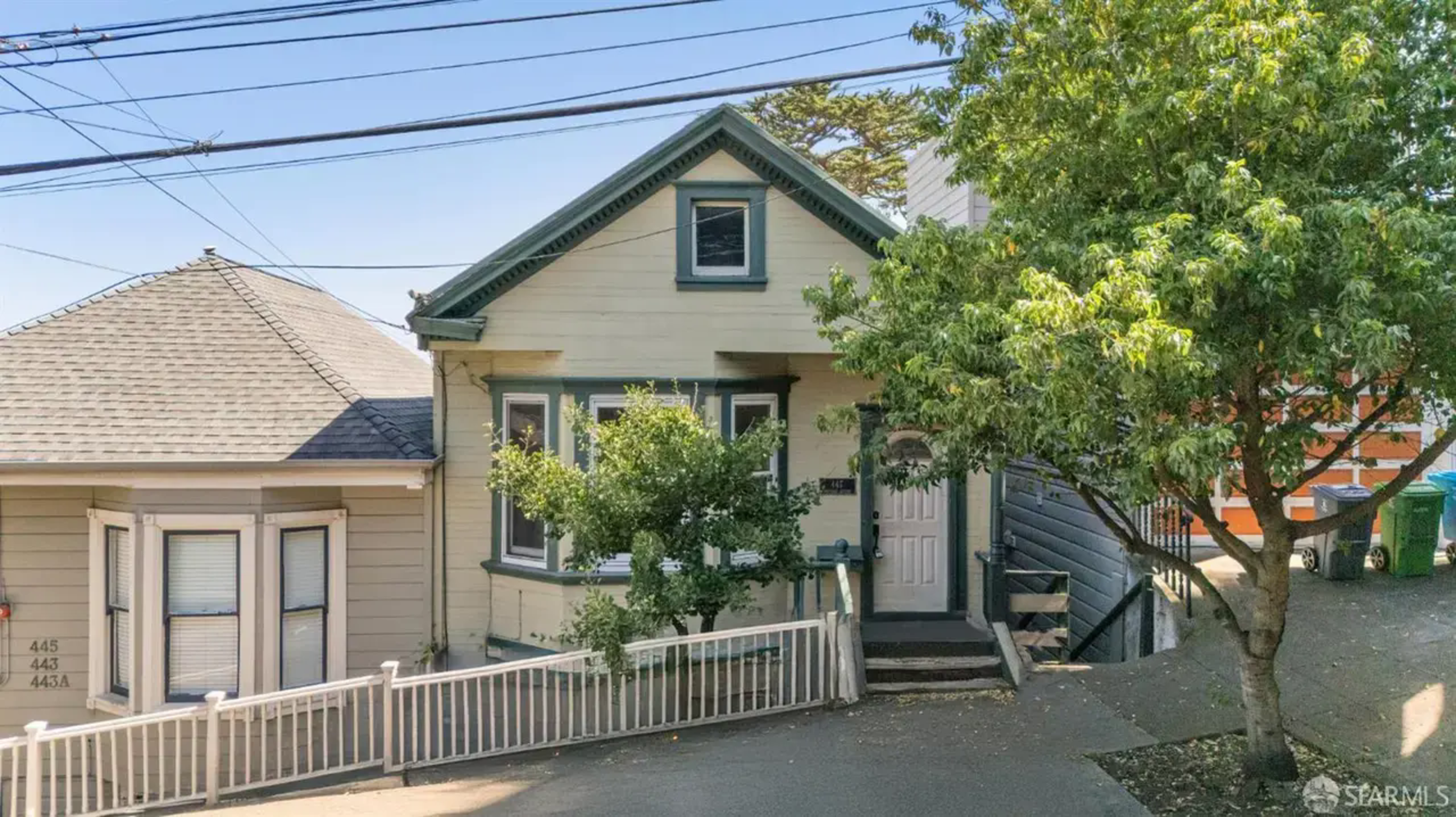 Two adjacent older homes sit on a sloped street, with one having a small tree in front and a white railing along the sidewalk.