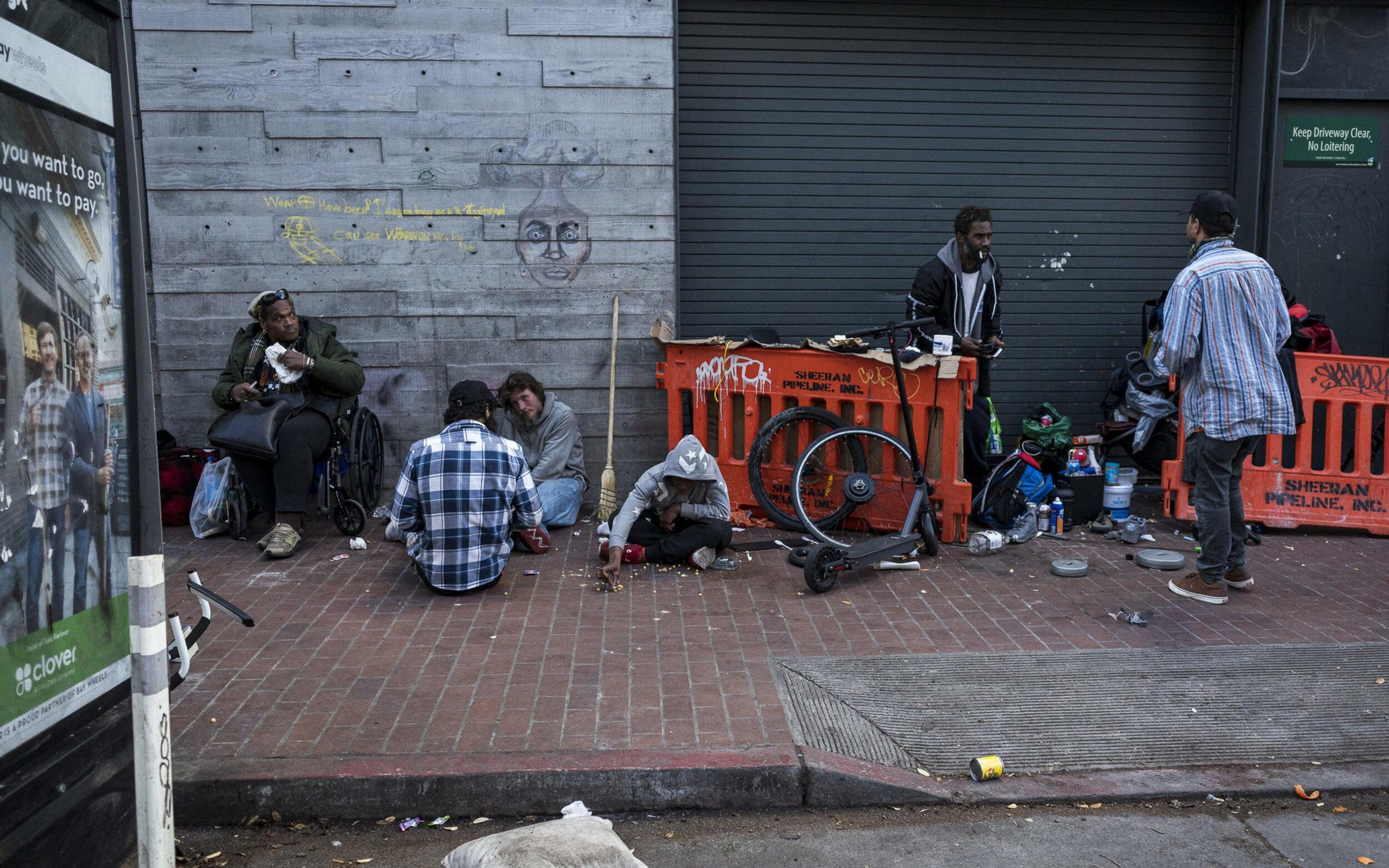 Several people sit and stand on a sidewalk beside a gray wall and orange barricades, with scattered belongings and a bicycle frame nearby.