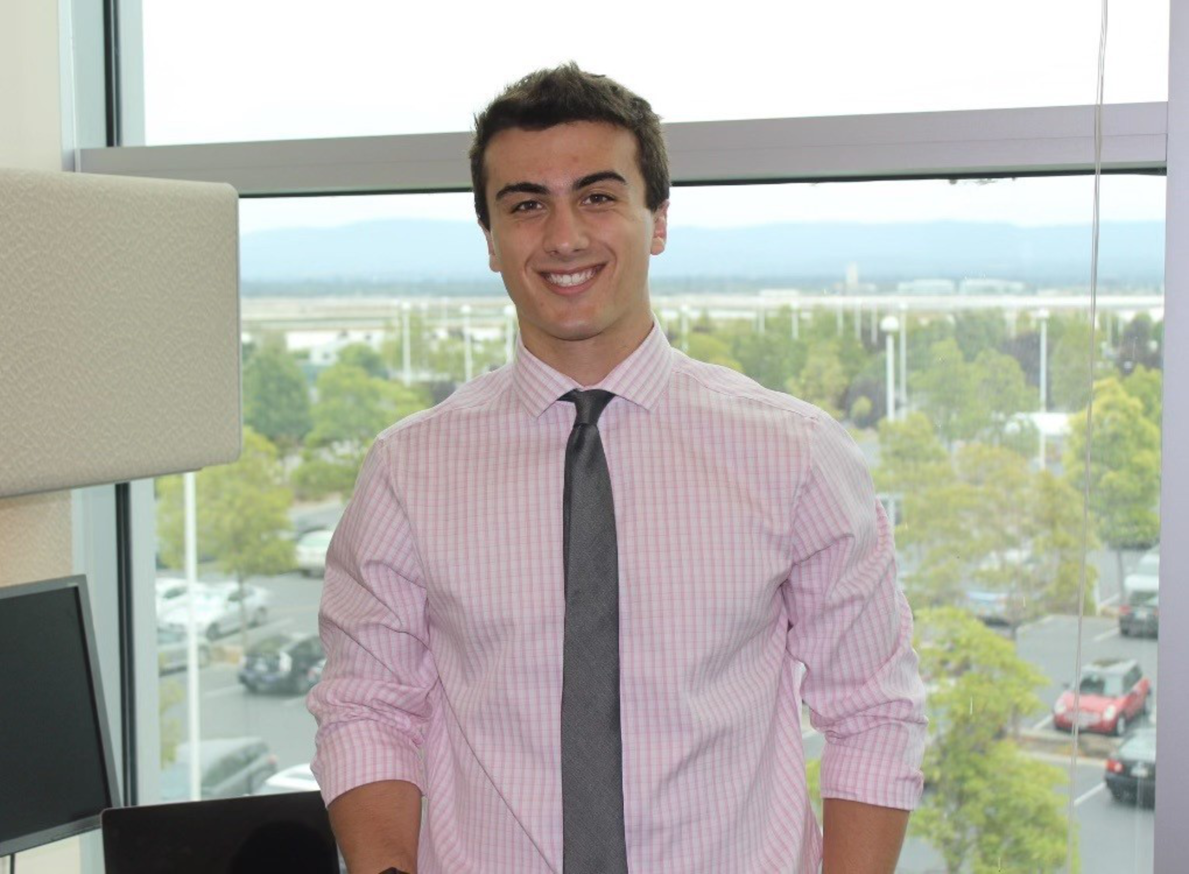 A young man in a pink checkered shirt and gray tie smiles while standing in front of a large window overlooking a parking lot and trees.