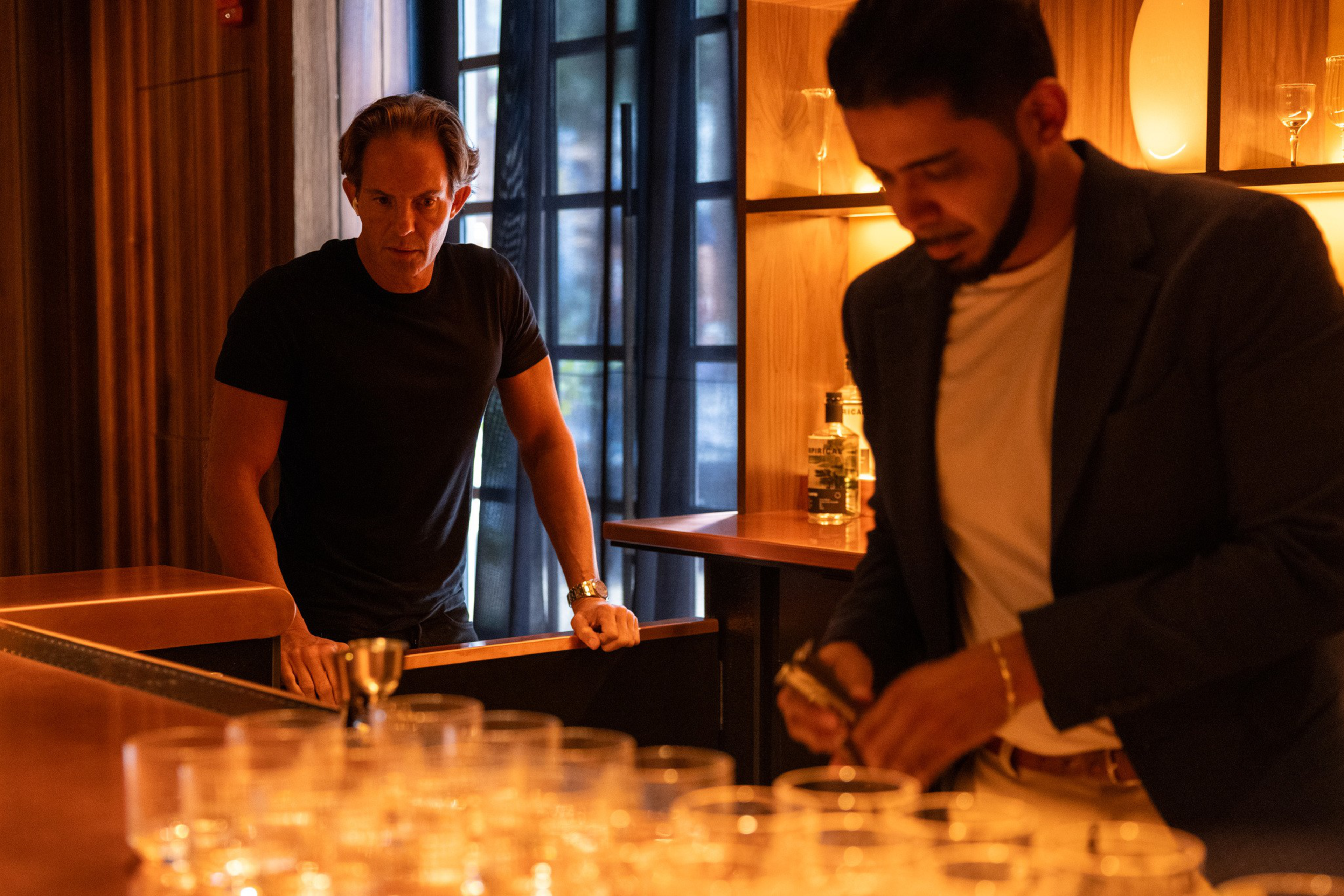 A man in a black shirt leans on a bar while another man in a blazer prepares drinks among rows of empty glasses in a warmly lit room.