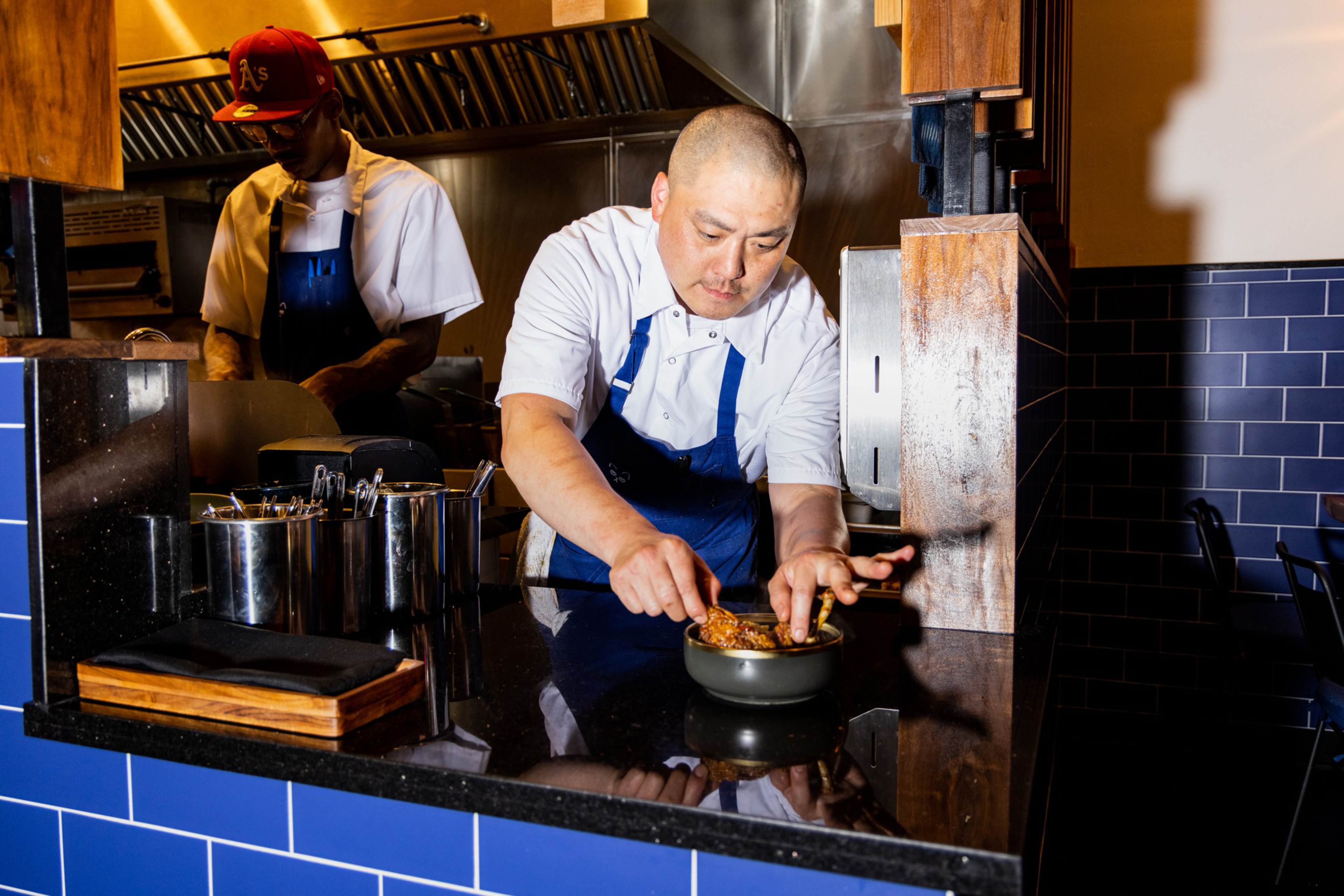 A chef in a white shirt and blue apron carefully places food in a bowl on a shiny black countertop, while another person cooks in the background.
