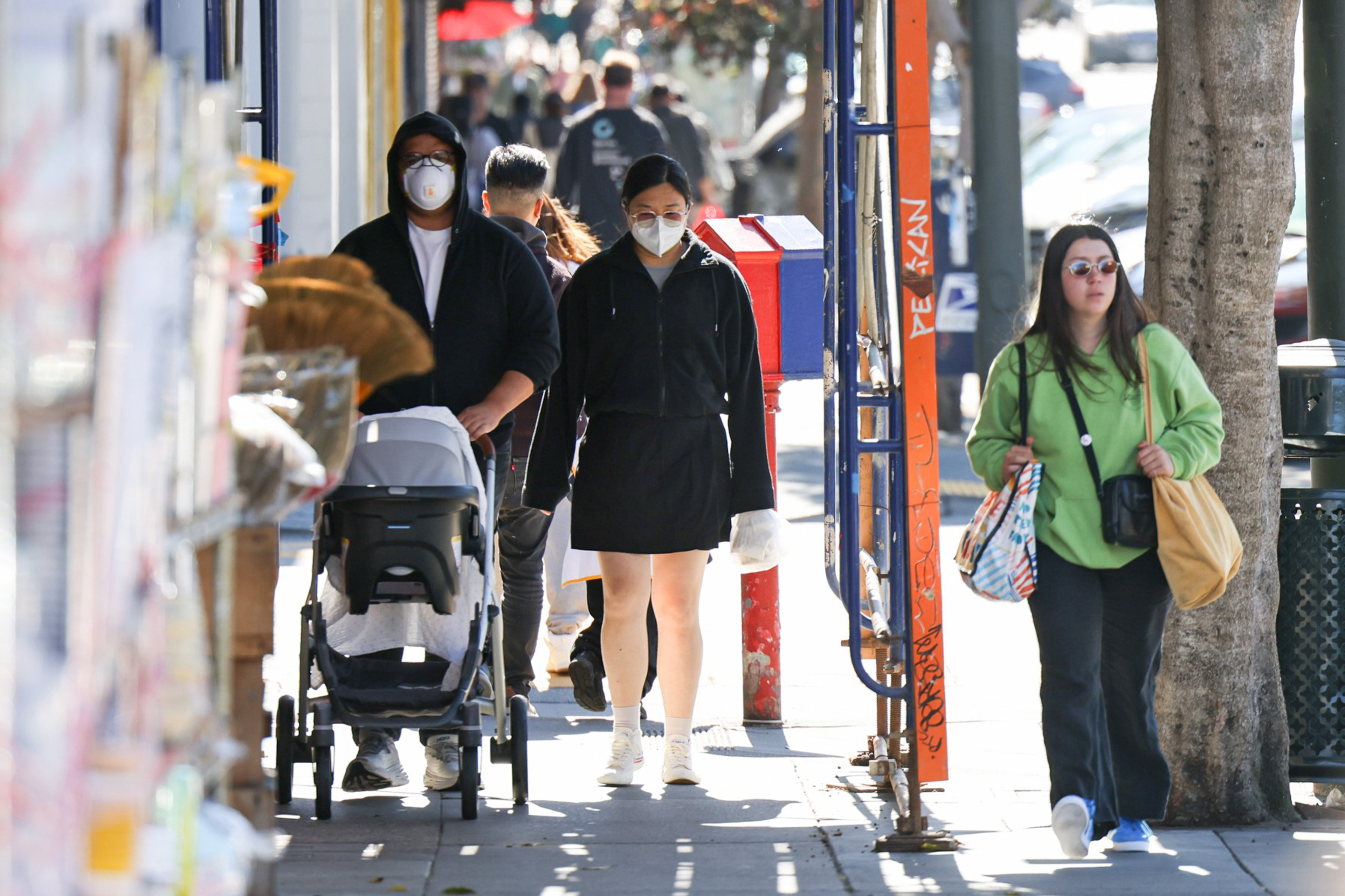 Several people walk on a sidewalk, two wearing face masks, one pushing a stroller, and another carrying two bags, near trees and a colorful mailbox.