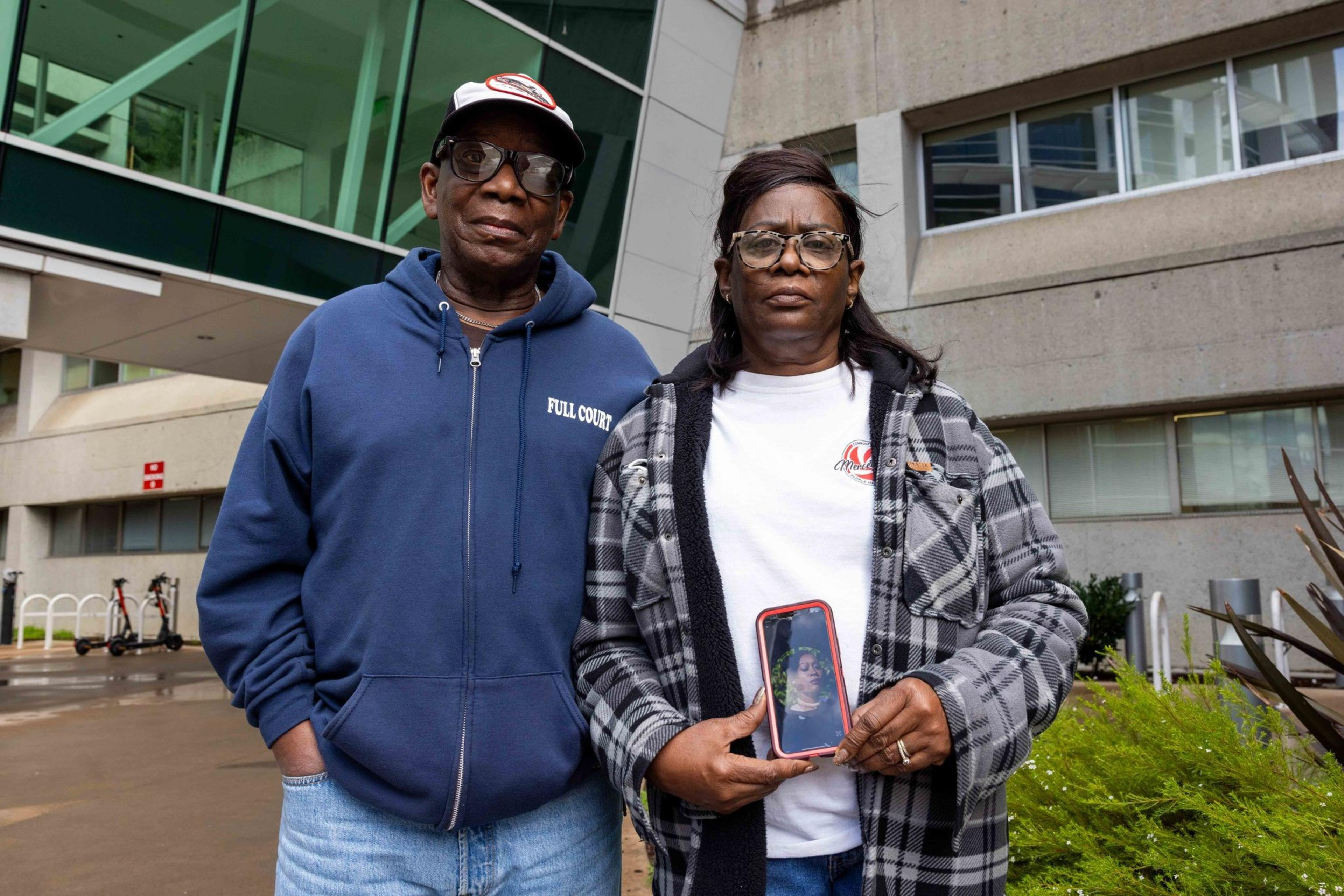 A man and woman stand outside a modern building. The man wears a blue hoodie and cap, while the woman, in a plaid jacket, holds a phone showing another person's photo.