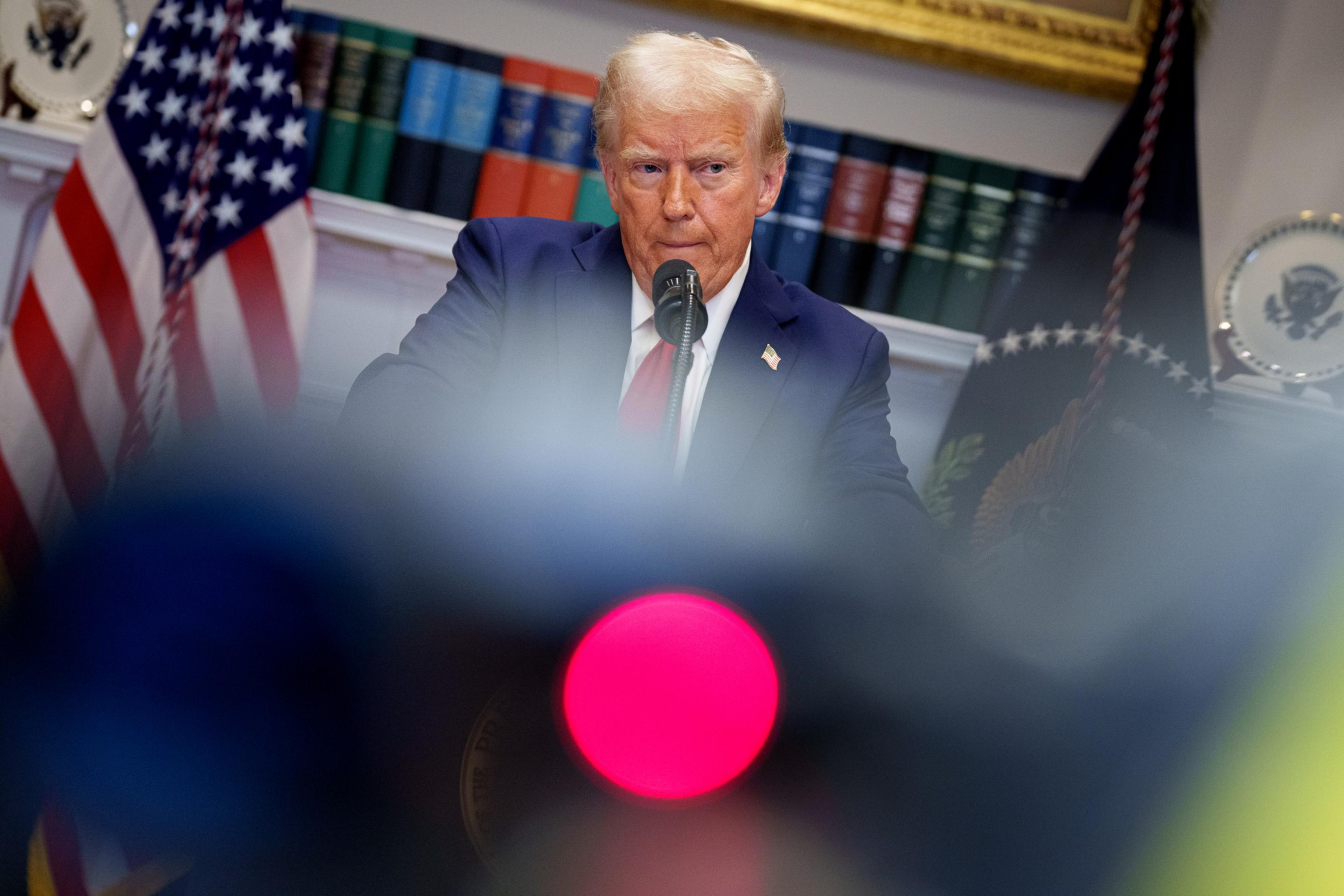 A man in a dark suit and red tie is speaking at a podium. Behind him are books, U.S. flags, and presidential emblems. The foreground is blurred with colorful lights.