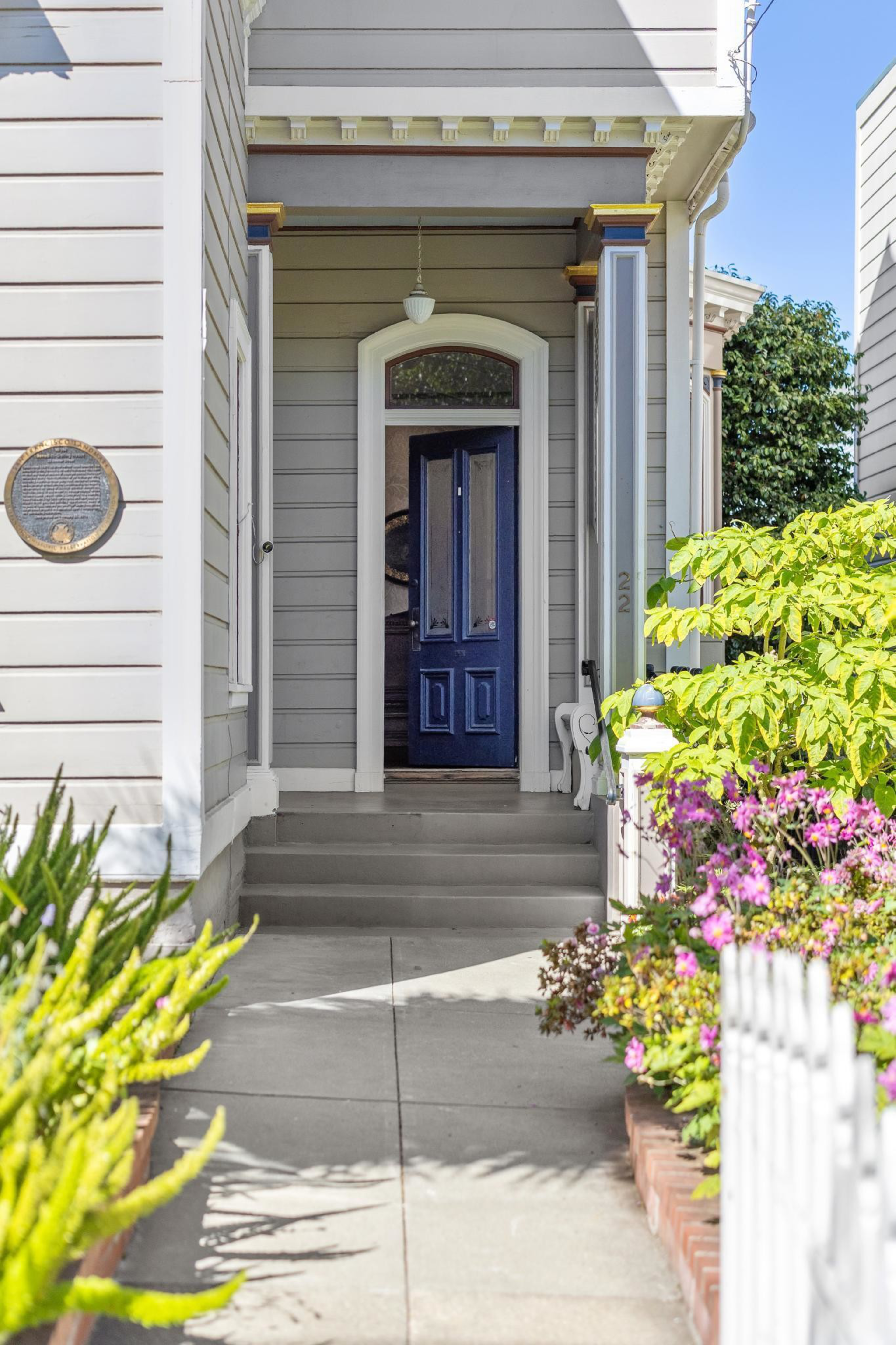 A walkway leads to a porch with gray siding, white trim, and a partially open dark blue door, surrounded by green plants and pink flowers.