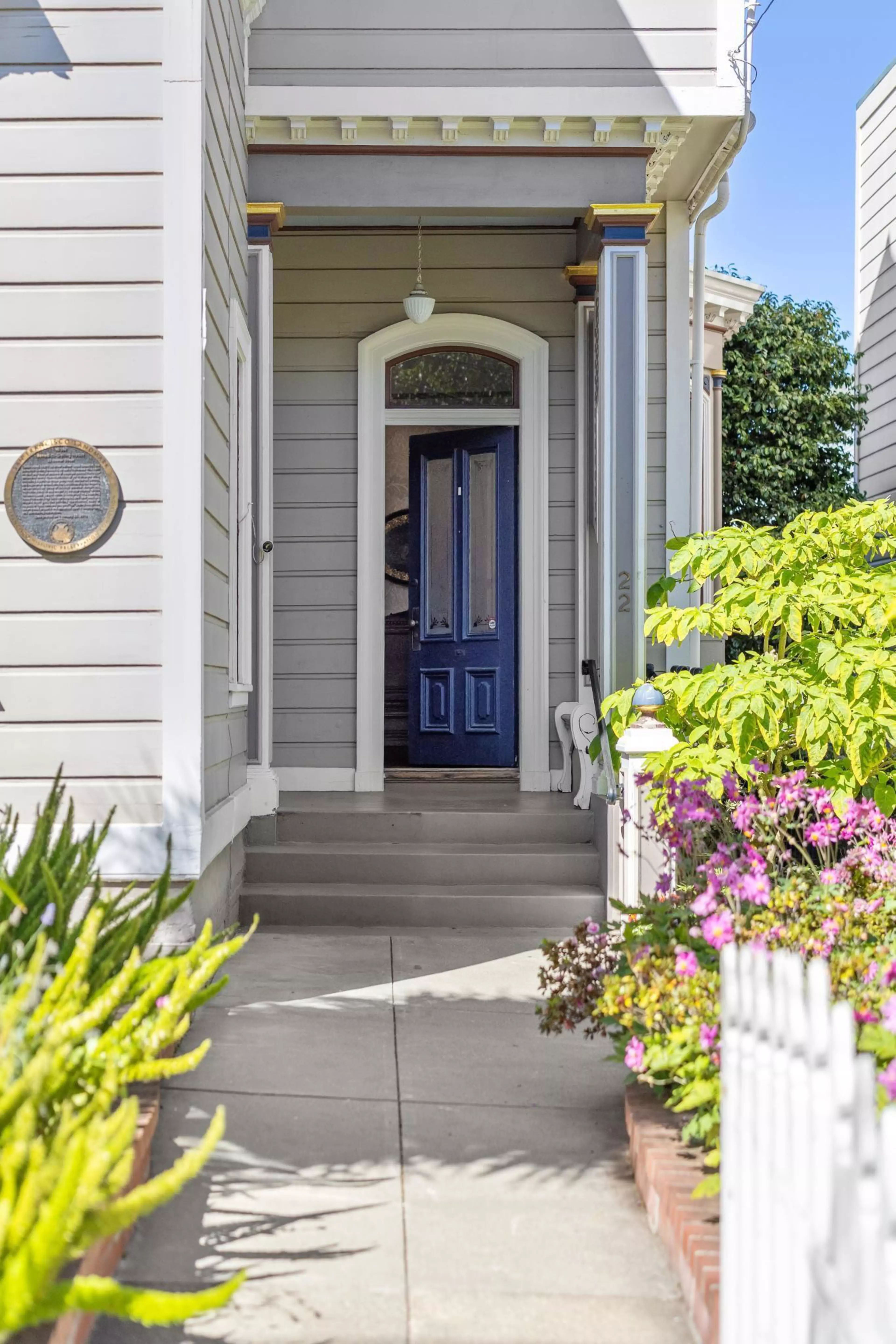 A walkway leads to a porch with gray siding, white trim, and a partially open dark blue door, surrounded by green plants and pink flowers.