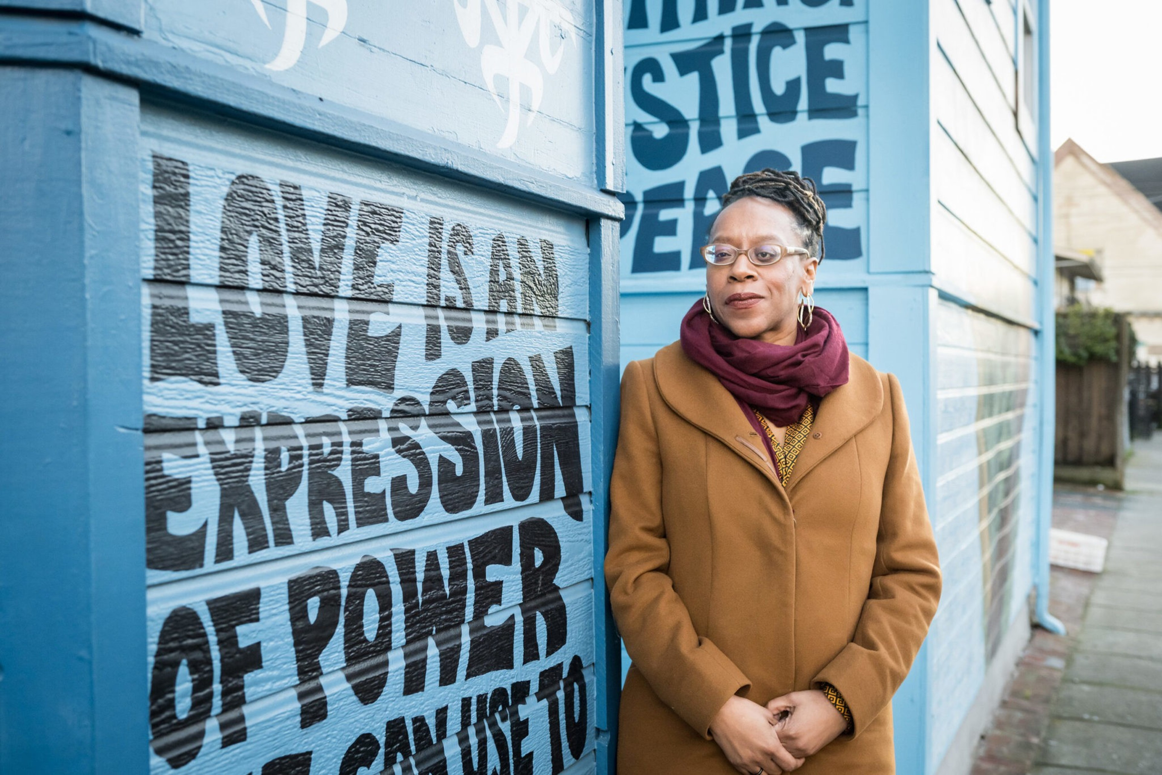 A woman wearing glasses, a brown coat, and a maroon scarf stands next to a blue wall with large bold text saying "Love is an expression of power."