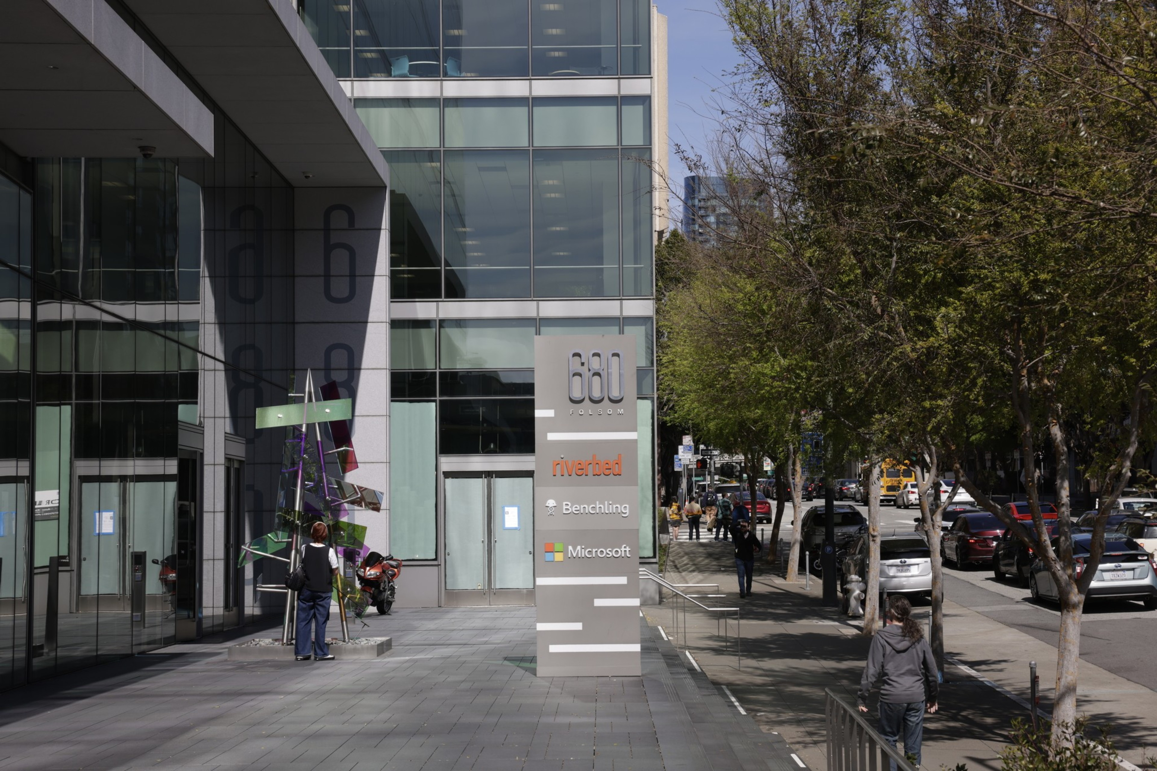 A city scene with a sign featuring company logos in front of a modern building, next to a sidewalk with trees and pedestrians.