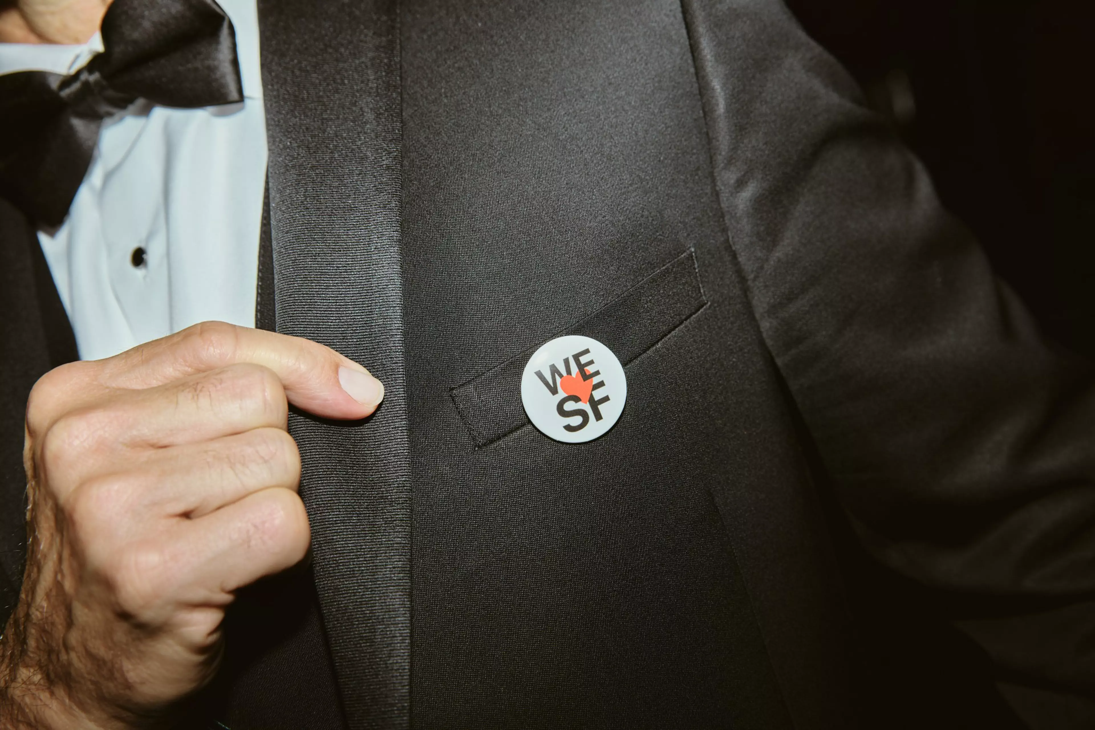 A person in a black tuxedo with a bow tie points to a round white pin on their jacket that says “WE SF” with a red heart.