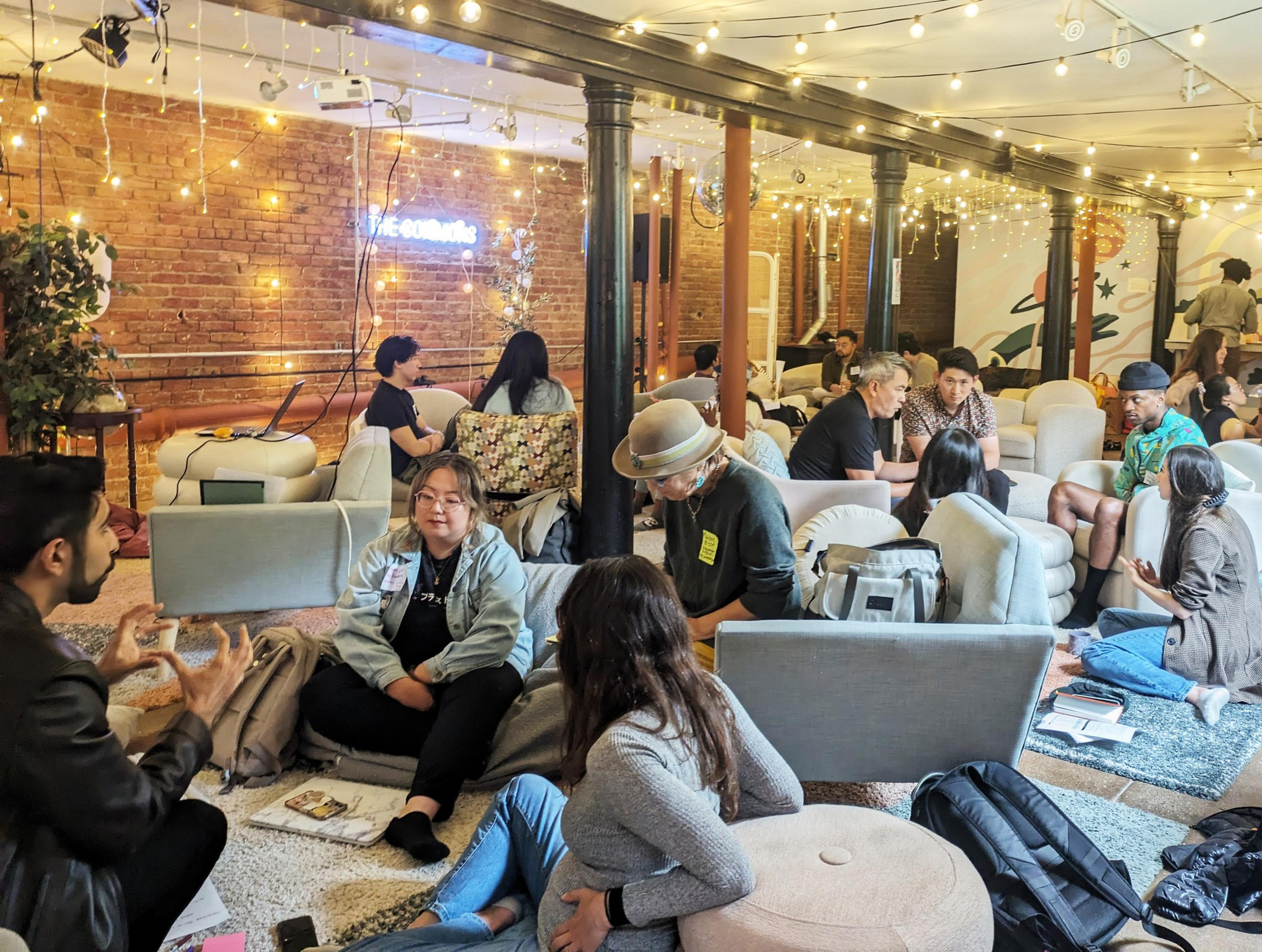 A cozy room with string lights and a brick wall backdrop. Groups of people are seated on sofas and cushions, engaged in discussions. A neon sign reads &quot;The something.&quot;