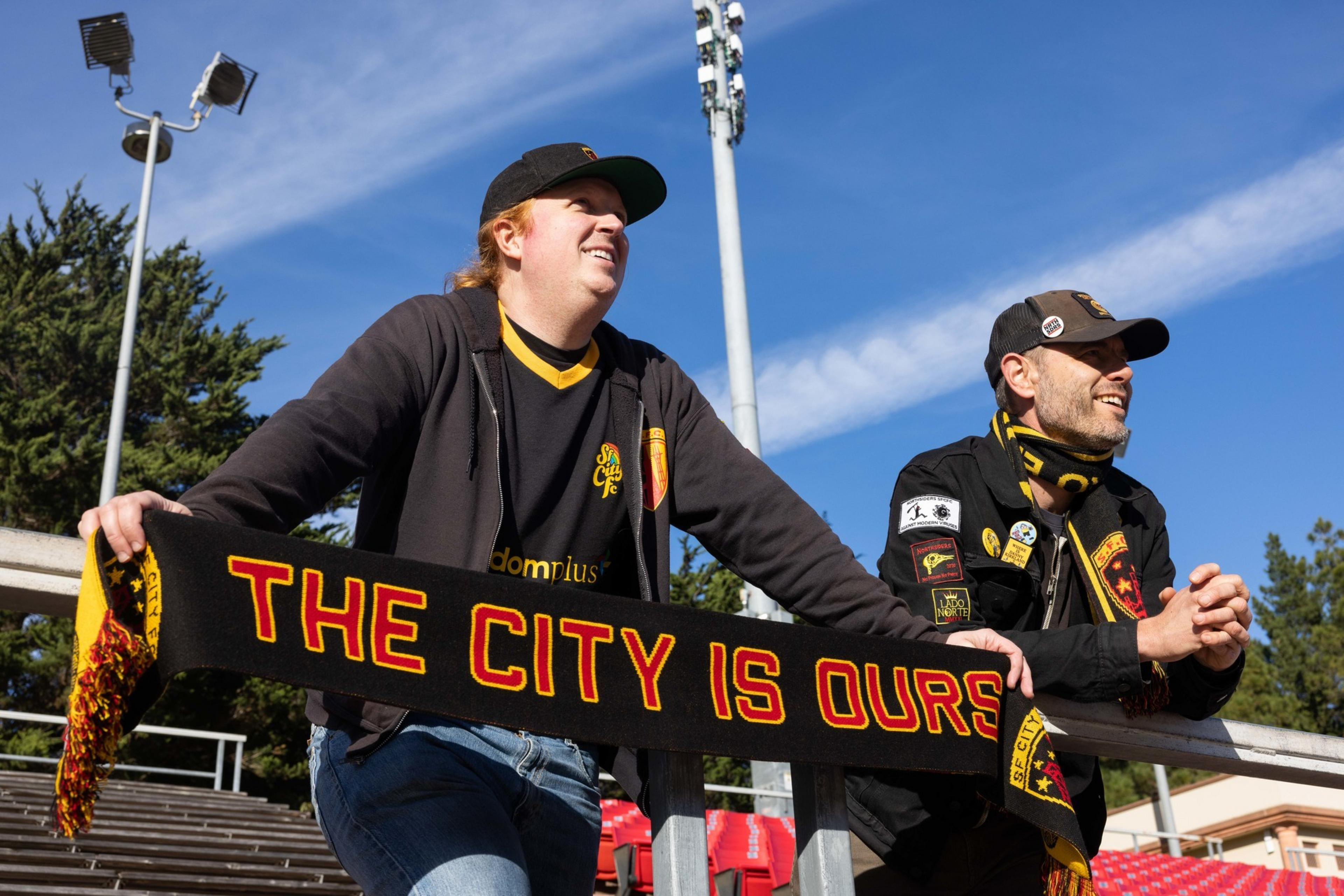 Two people in jackets and caps stand in a stadium holding a scarf that reads &quot;THE CITY IS OURS,&quot; with trees and floodlights in the background.