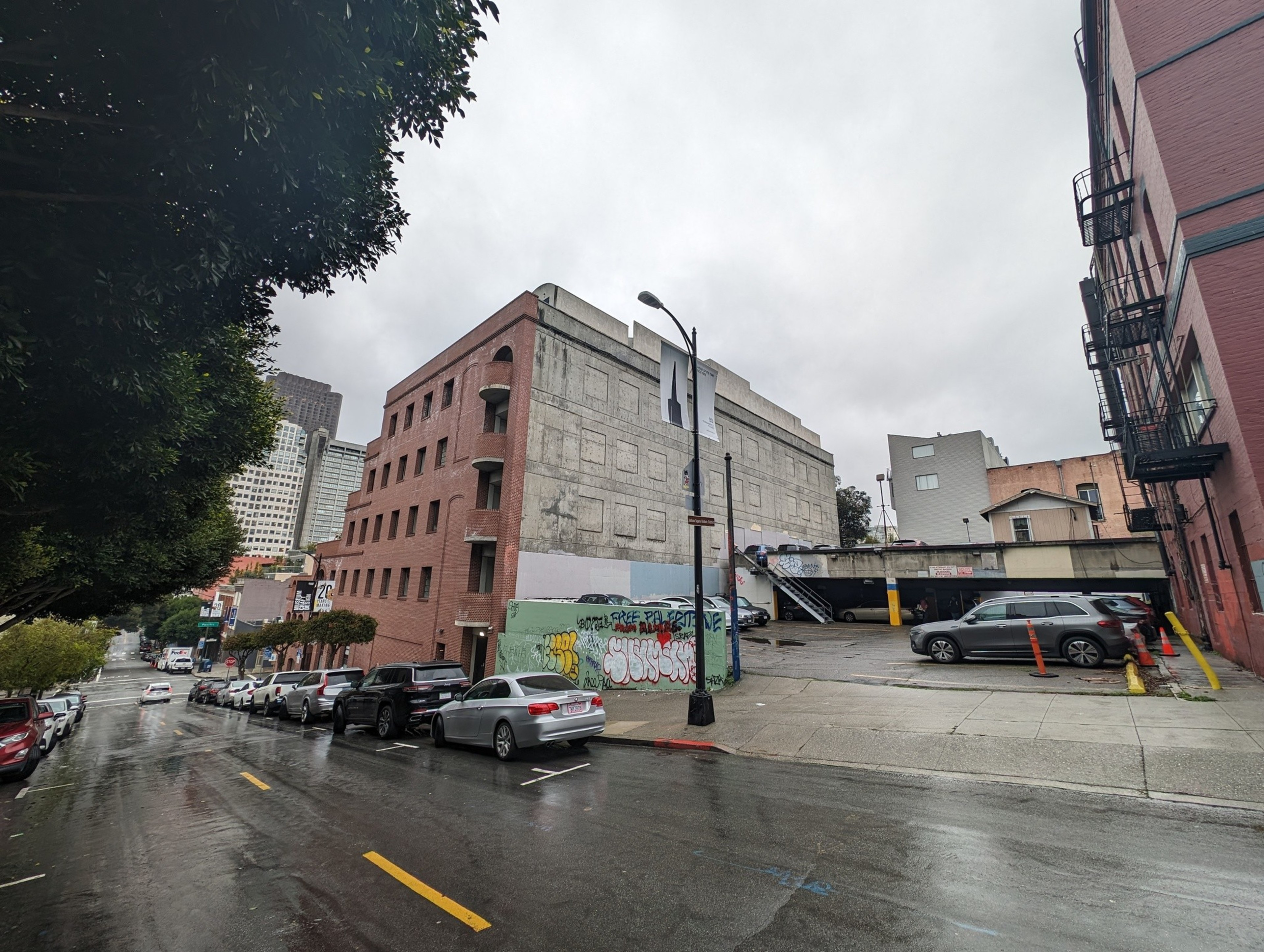 A bi-level street parking lot sits between two brick-exterior buildings on a cloudy rainy weekday afternoon.