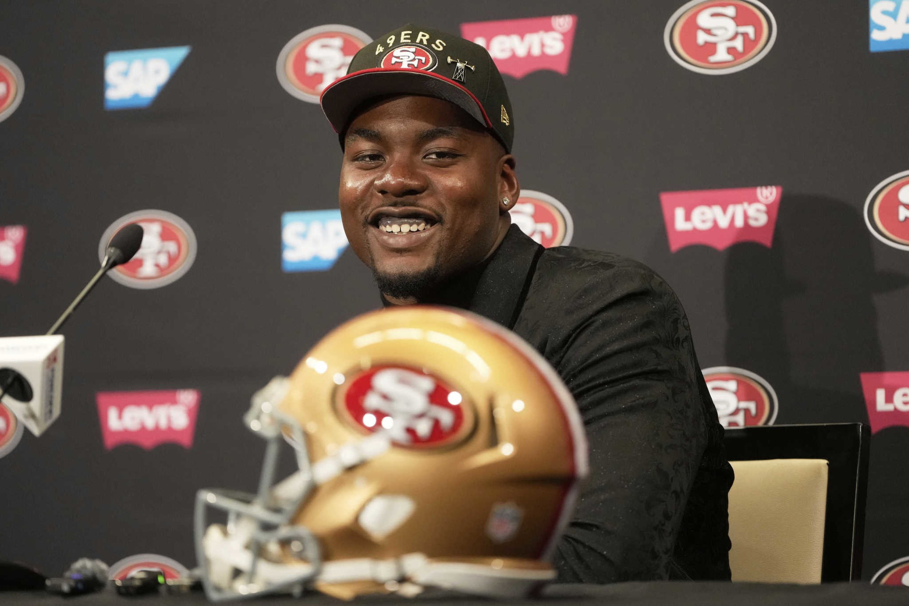 A person wearing a 49ers cap is smiling at a press conference. A microphone is in front of them, and a 49ers football helmet is on the table. Logos are in the background.