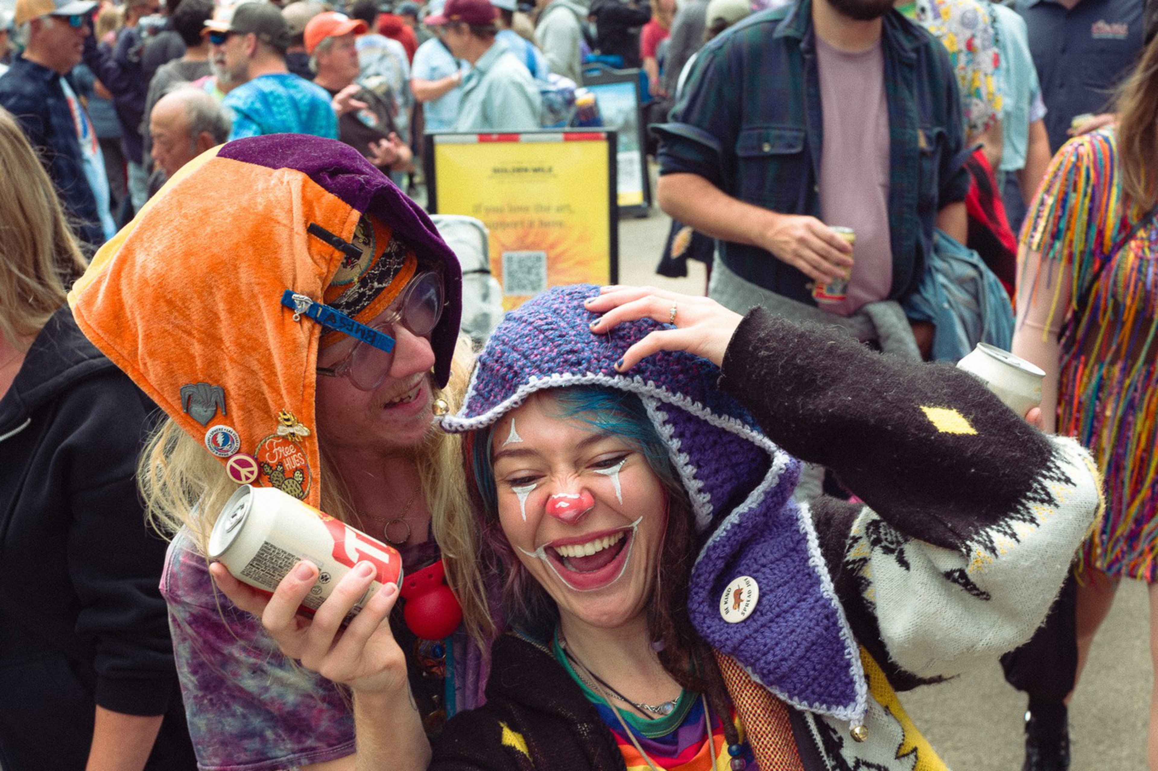 Two people wearing colorful, playful outfits and hats laugh while taking a selfie at an outdoor event with a crowd in the background.