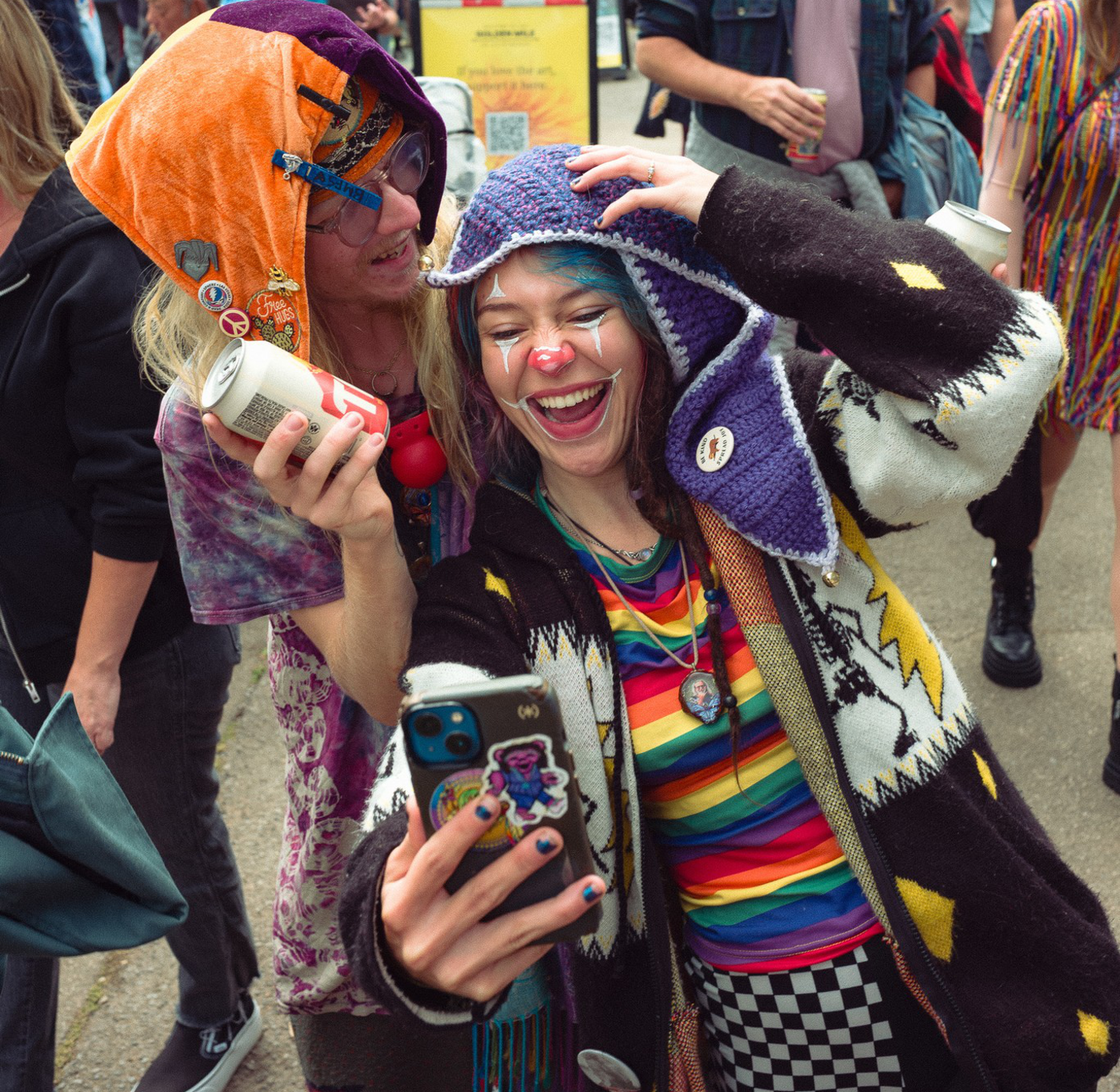 Two people wearing colorful, playful outfits and hats laugh while taking a selfie at an outdoor event with a crowd in the background.