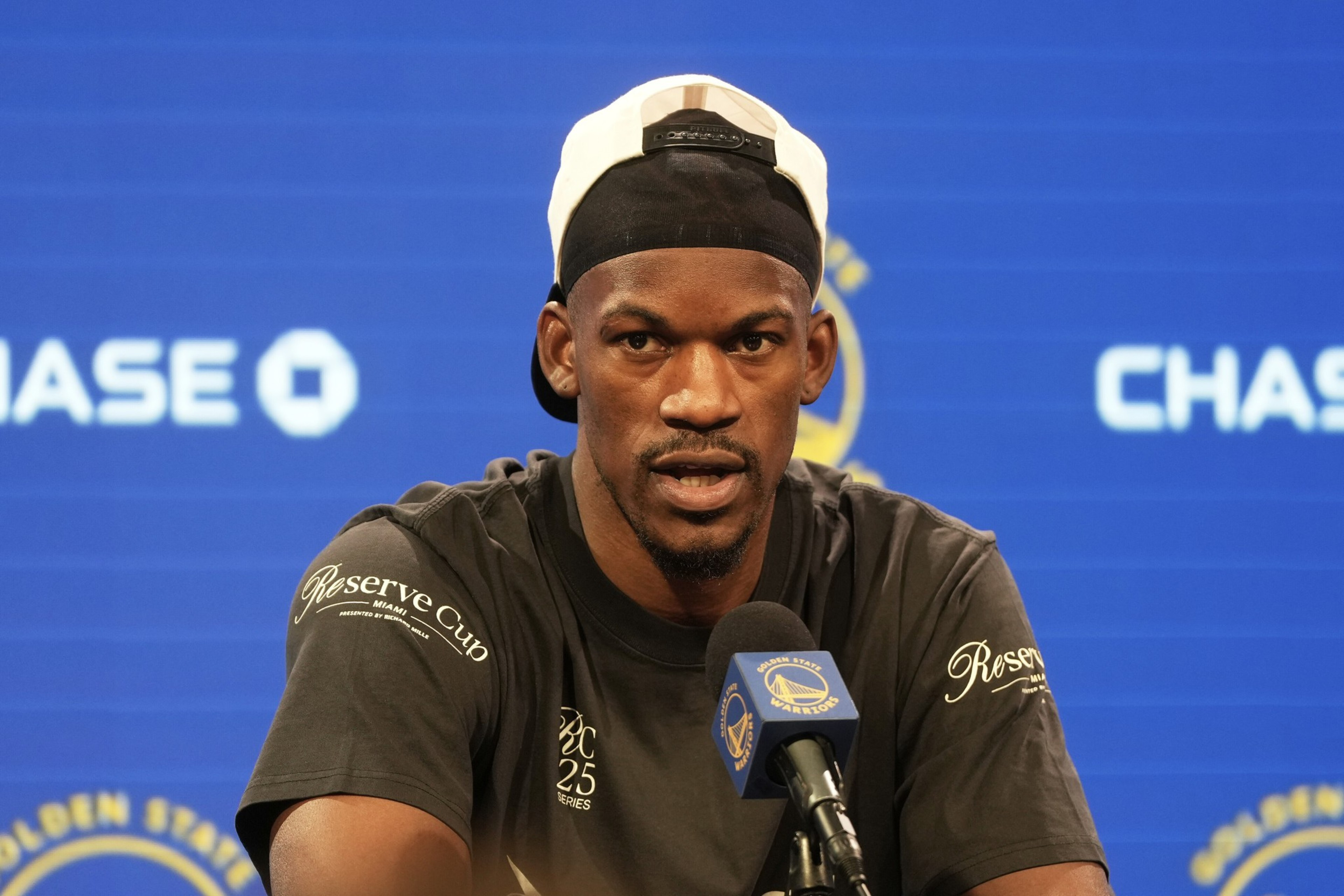 A man wearing a dark shirt and backwards cap sits at a press conference table with Golden State Warriors and Chase logos on a blue backdrop behind him.