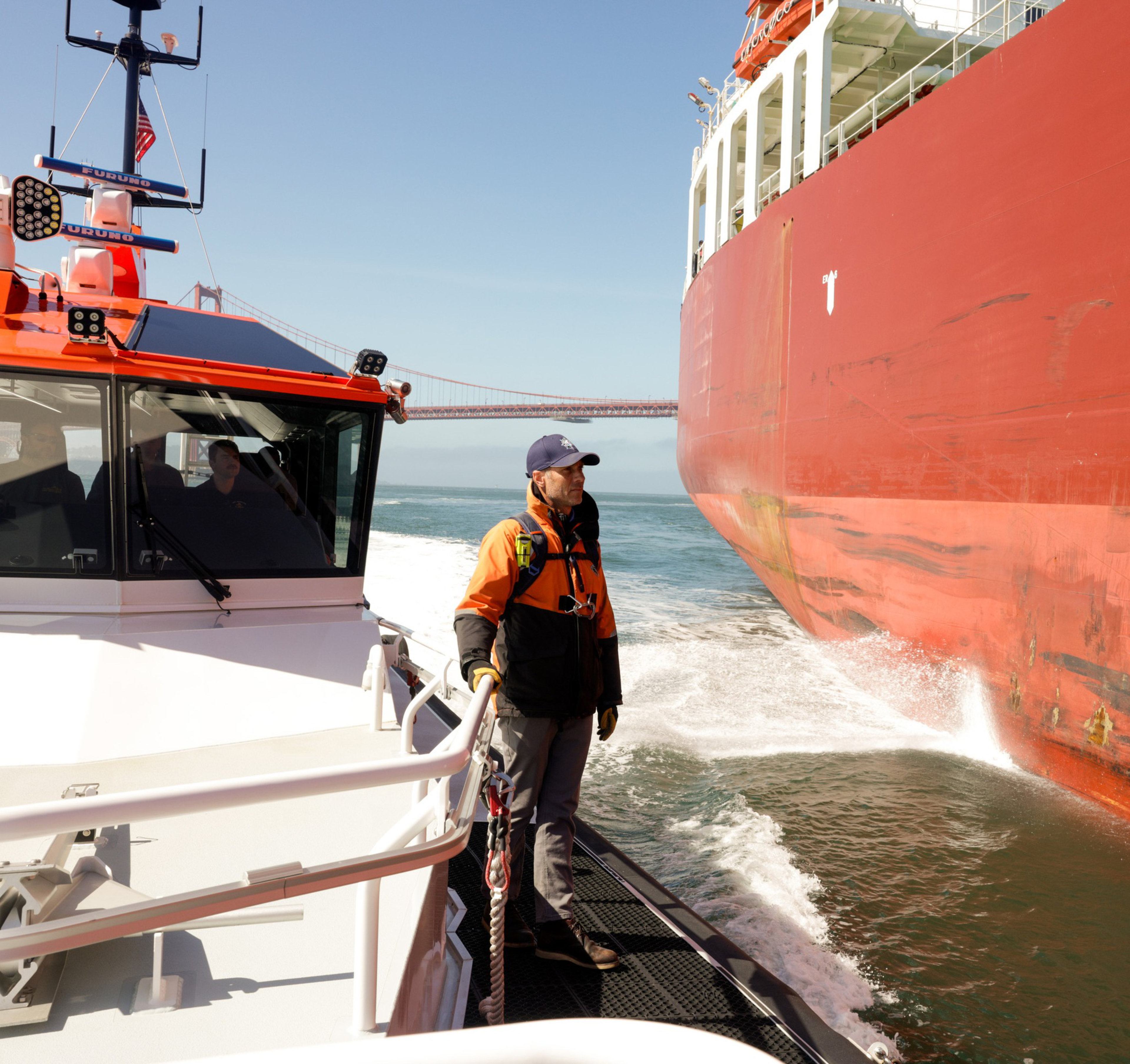 A person stands on a small boat beside a large red ship, with a bridge in the background.