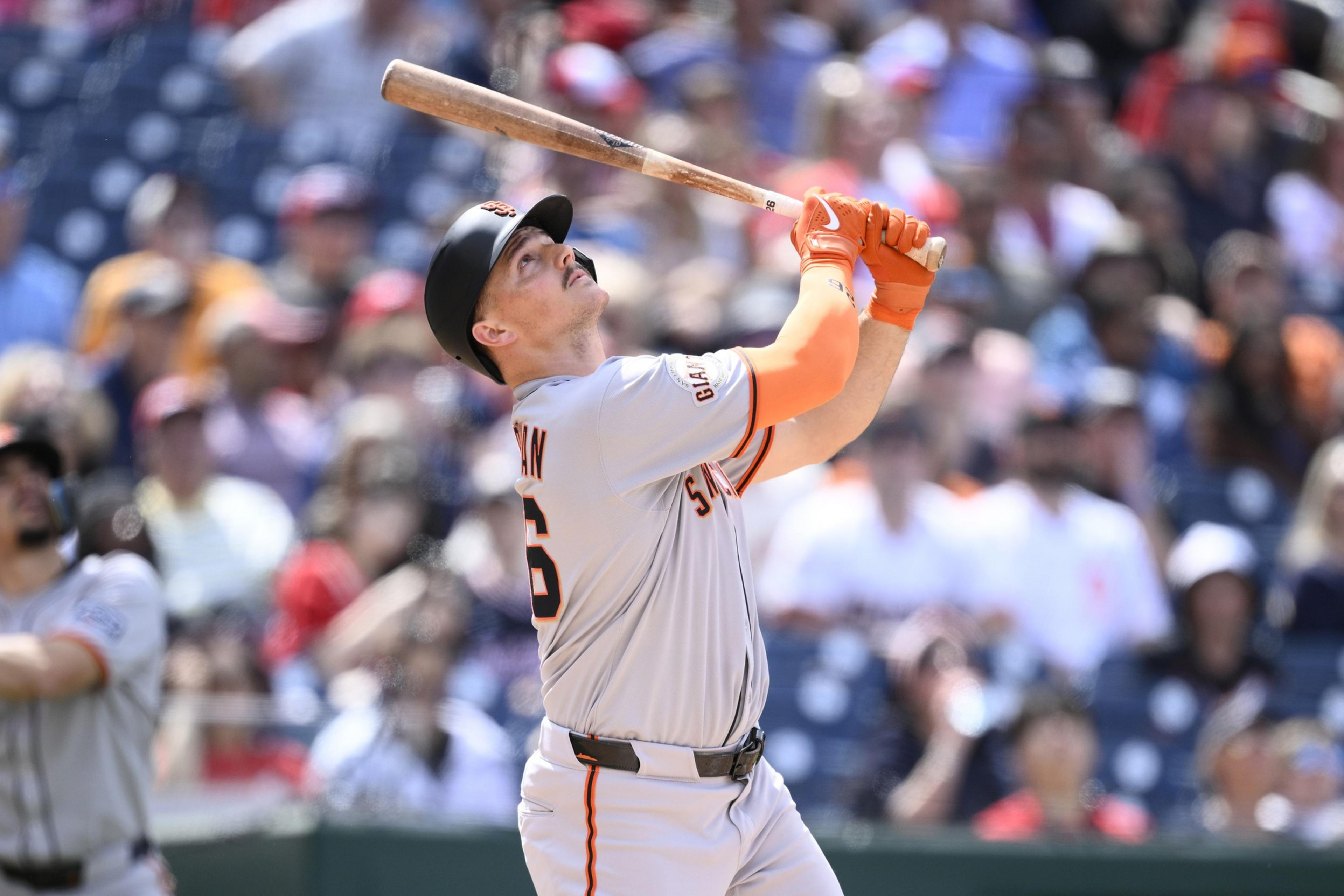 A baseball player in a gray uniform and black helmet watches his hit while holding a bat. The crowd is blurred in the background, suggesting a sunny day at a game.