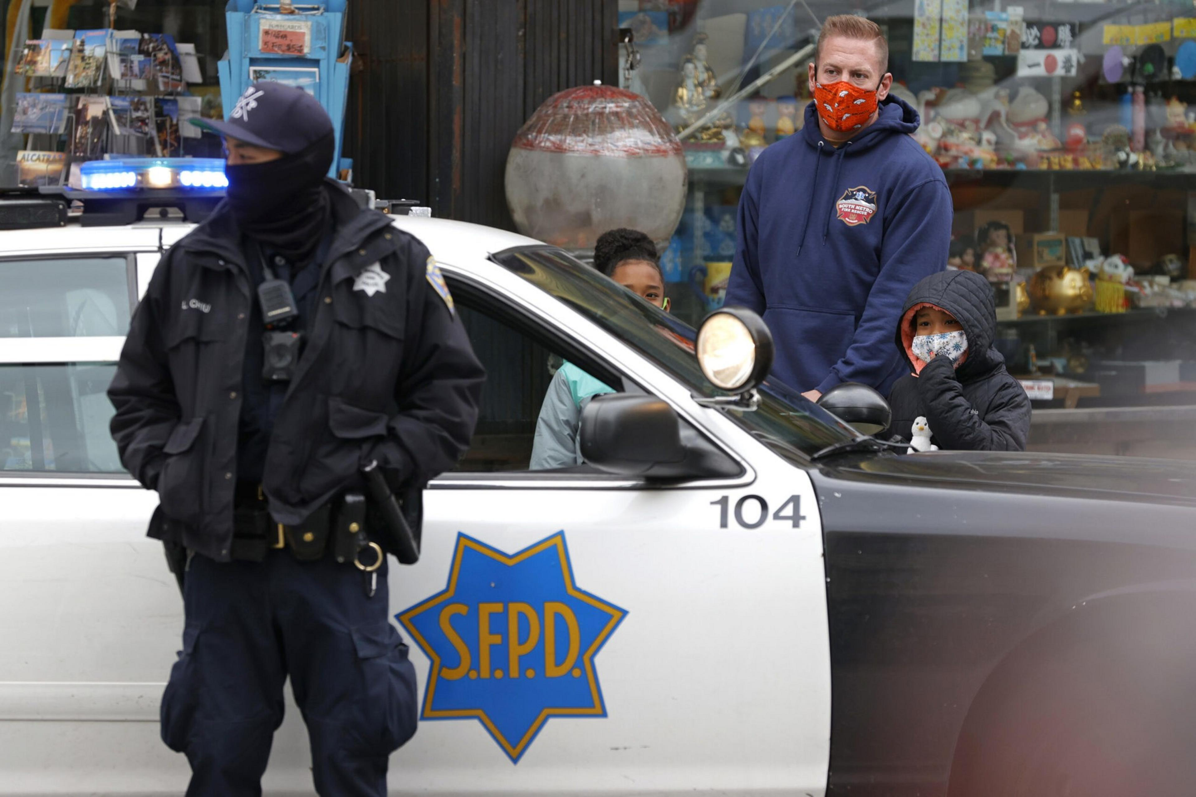 A San Francisco police officer in dark uniform stands beside a police car while three masked people, including a child, watch from behind.