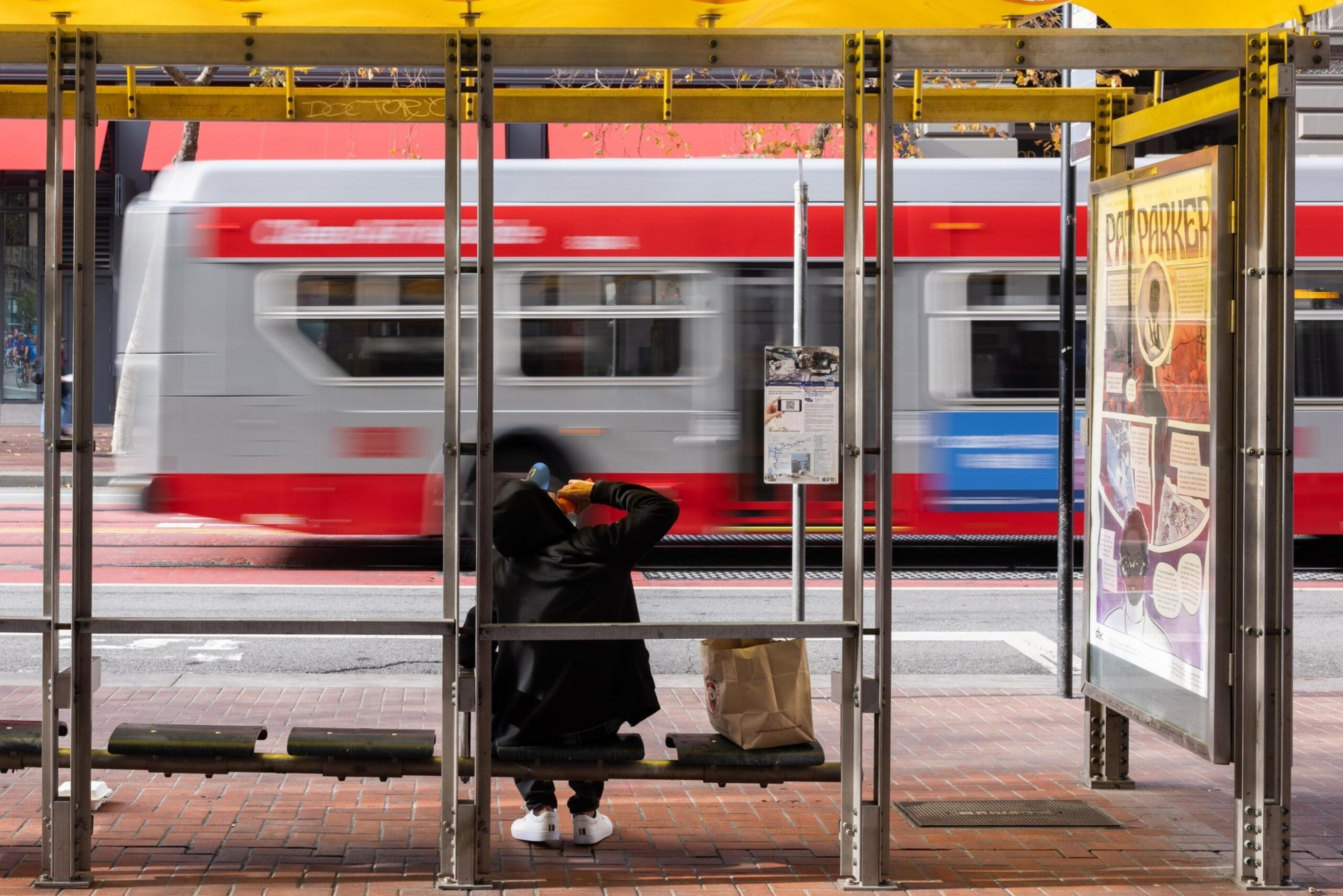 A person in a black hoodie and white shoes sits at a bus stop, drinking from a can as a red and silver bus speeds by behind them.