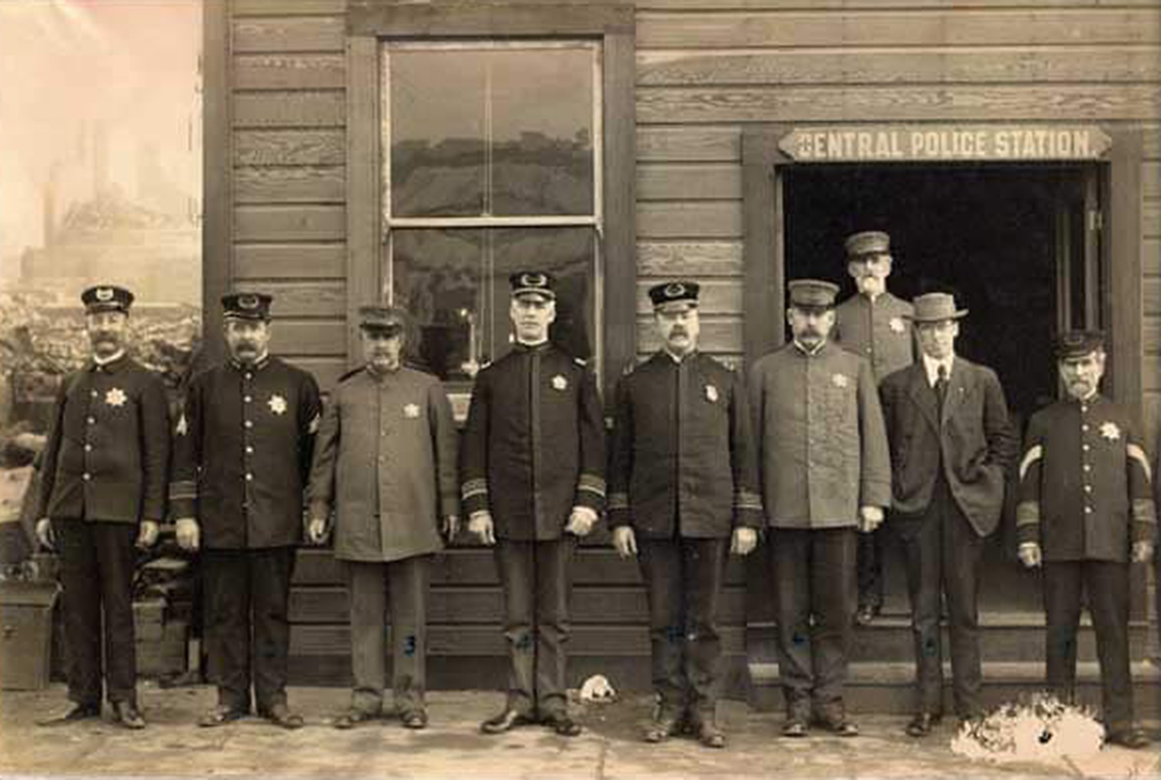 Nine men, mostly in early 20th-century police uniforms, stand and pose outside a wooden building labeled "Central Police Station."