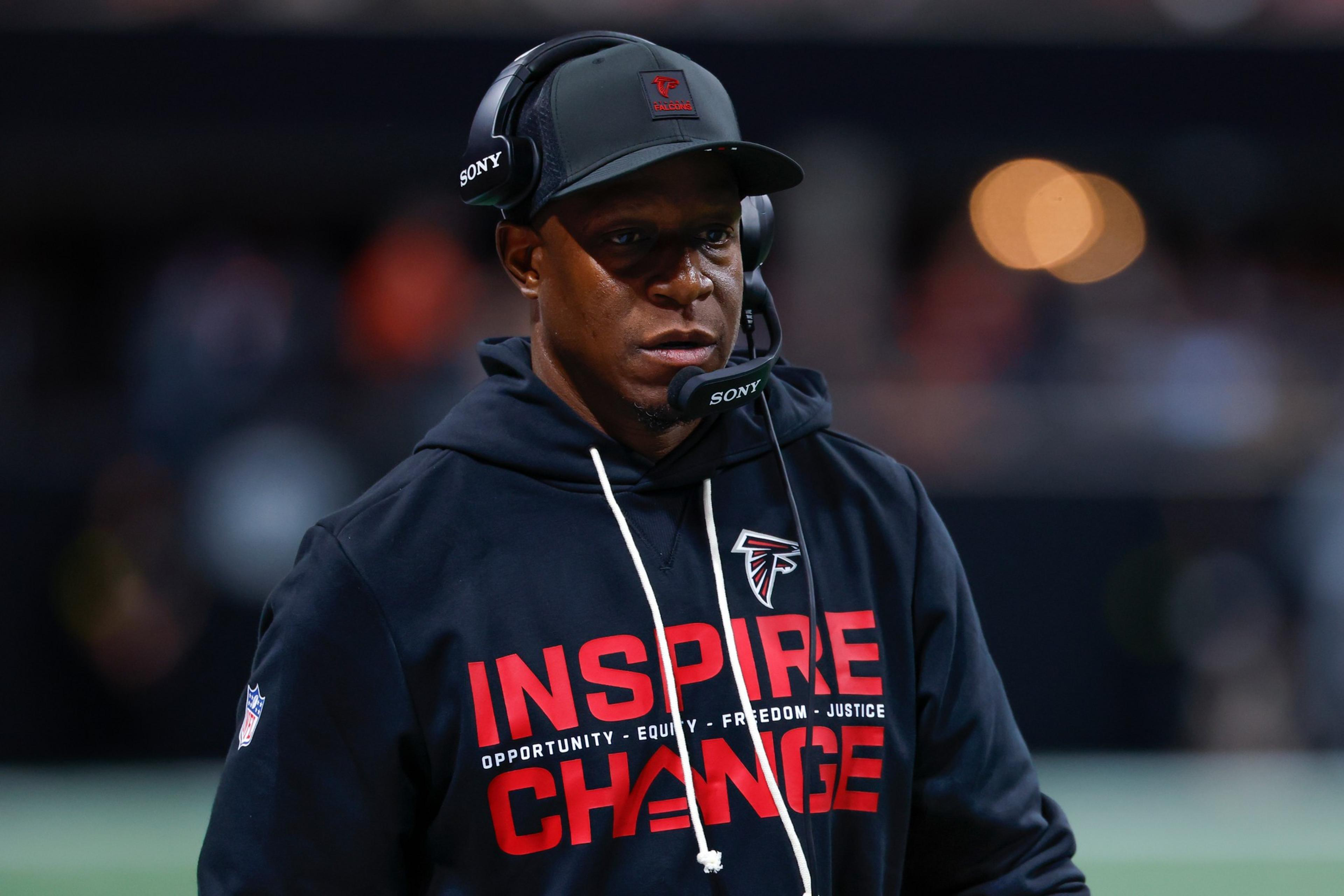 A man wears a black hoodie with “INSPIRE CHANGE” in red letters and a Falcons logo, along with a headset and cap, focused during a sports event.