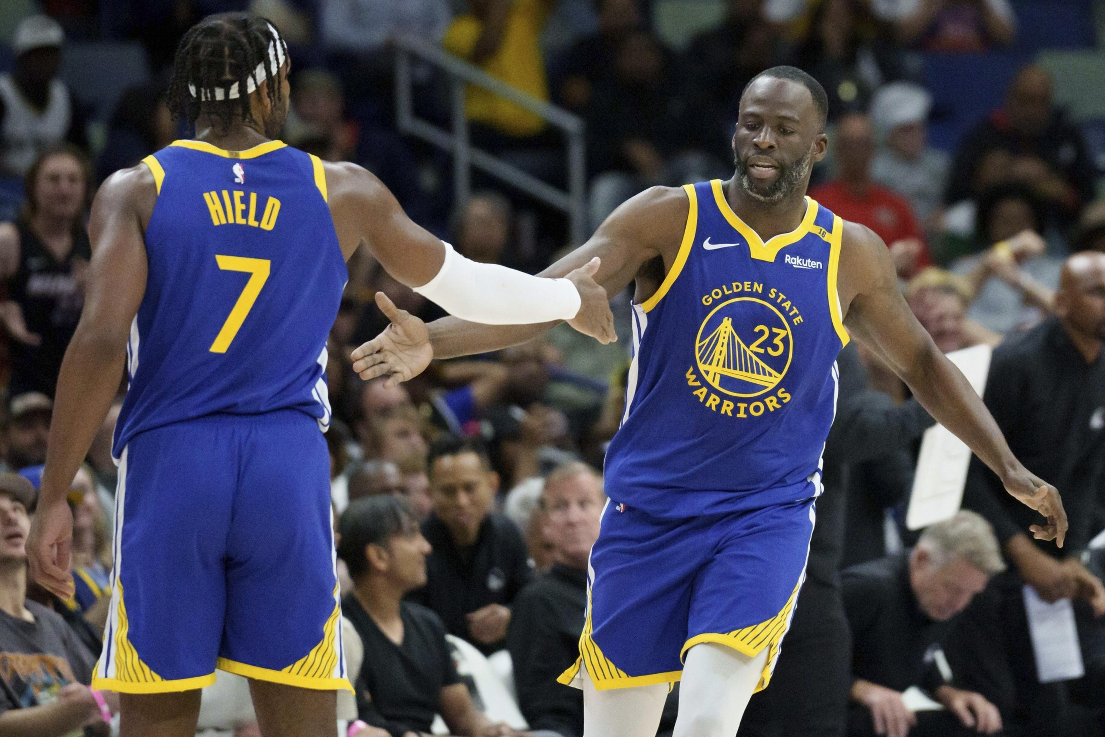 Two basketball players in blue Golden State Warriors jerseys high-five on the court. One wears number 7, the other 23. The crowd is visible in the background.