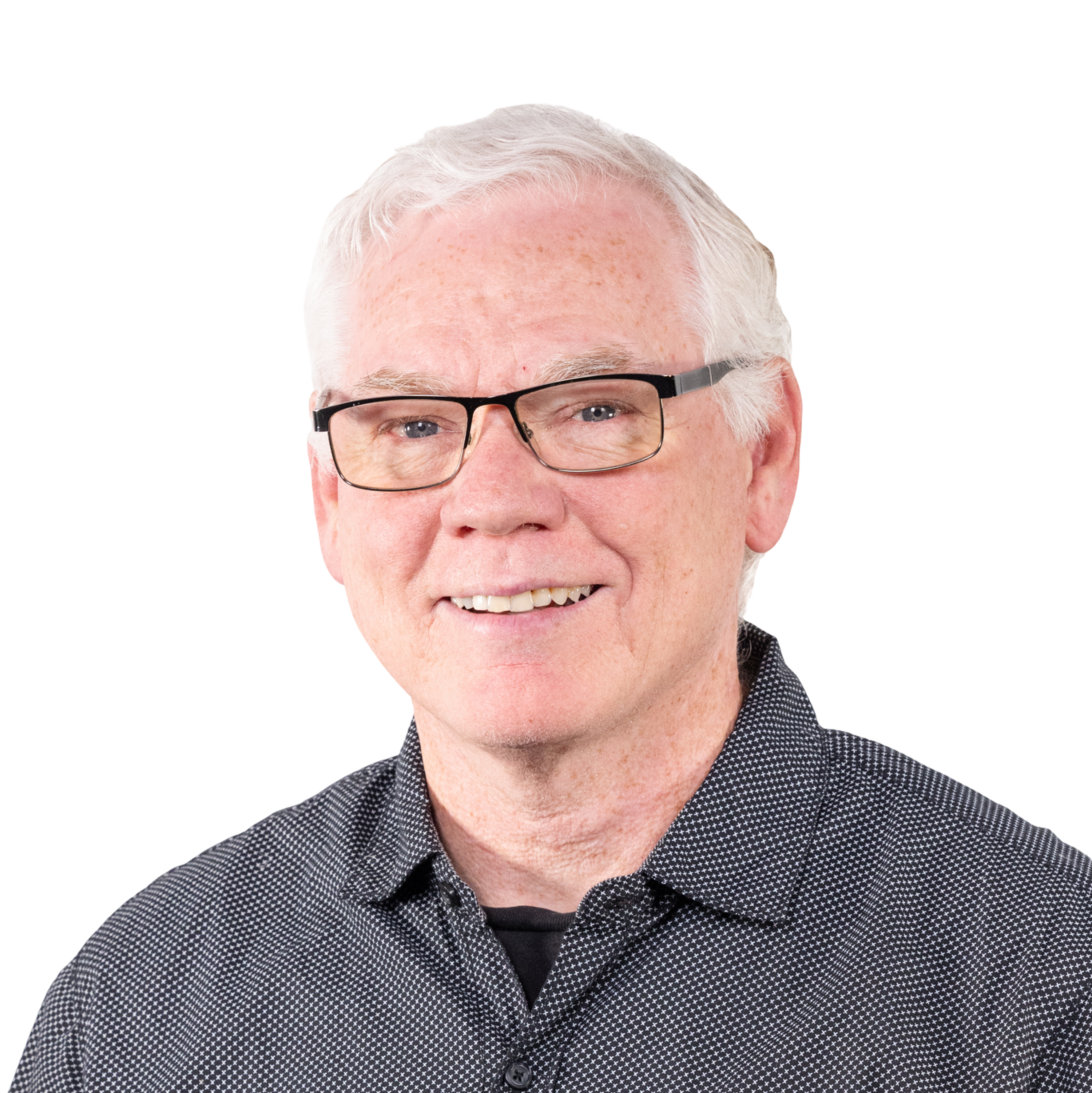 An older man with white hair and glasses smiles while wearing a dark, patterned button-up shirt over a black undershirt.