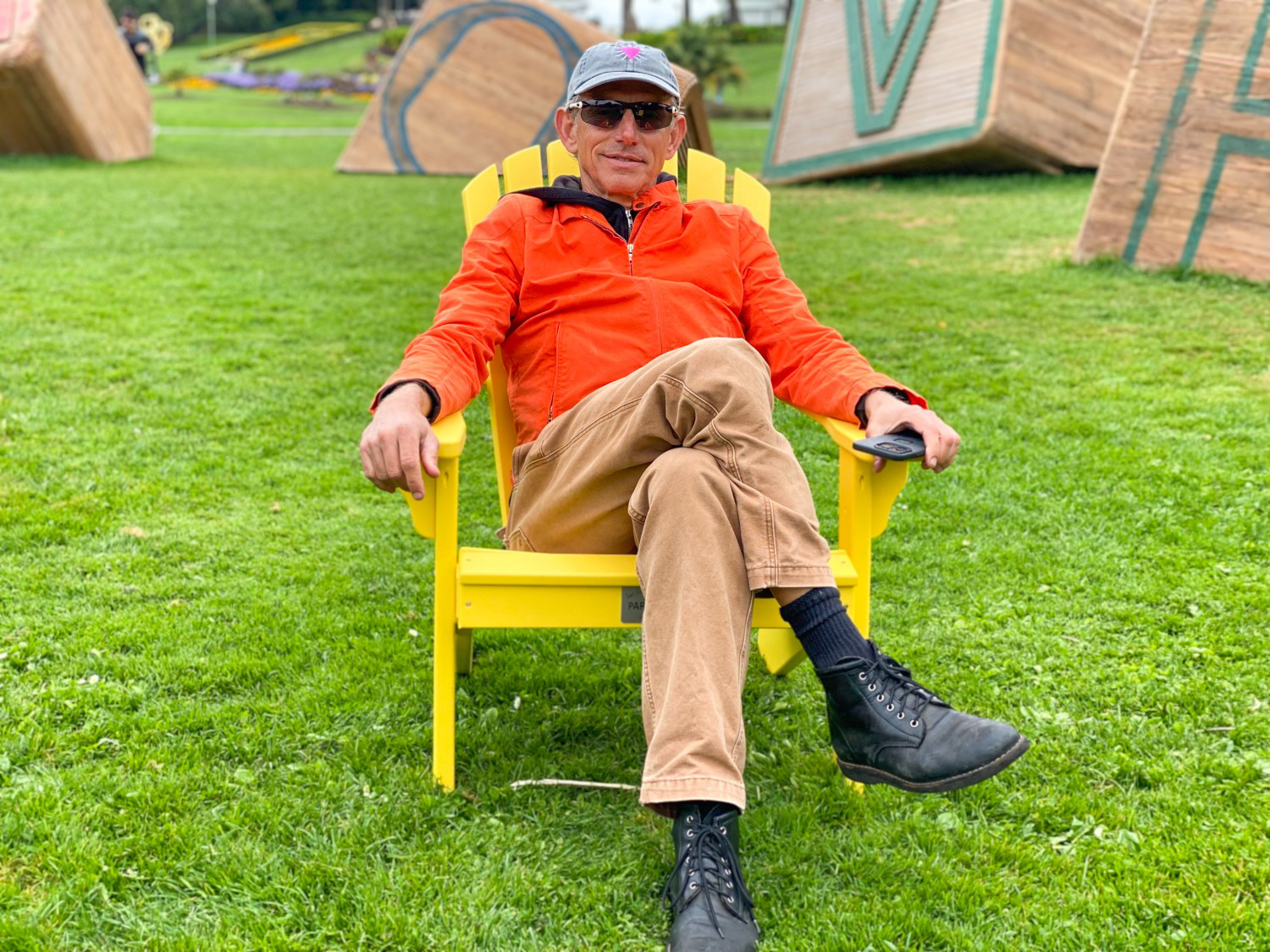 A man wearing an orange jacket, beige pants, black boots, and sunglasses sits relaxed in a bright yellow chair on green grass.