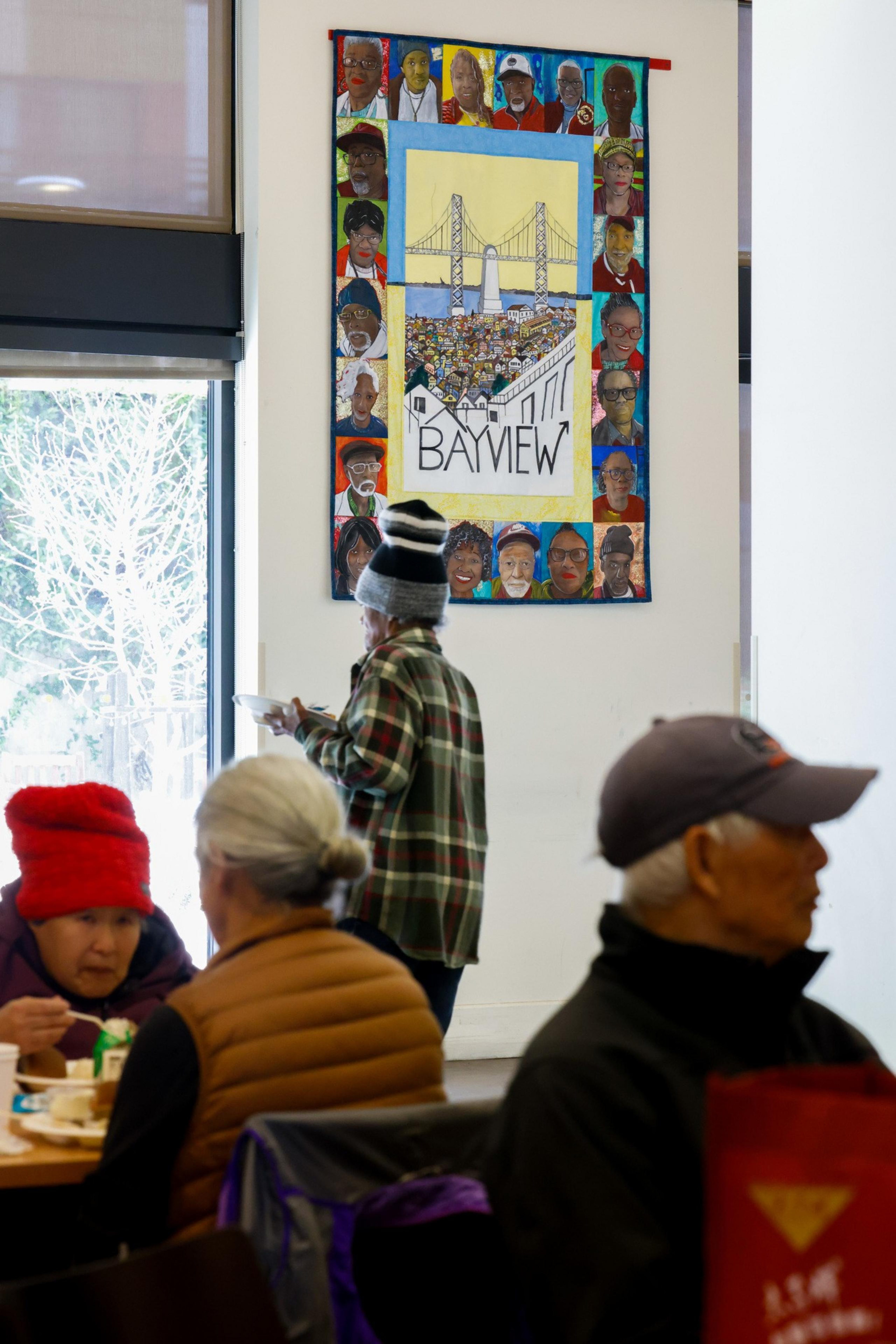 The image shows people sitting and eating in a room with a colorful mural on the wall depicting faces and a bridge labeled &quot;BAYVIEW.&quot;