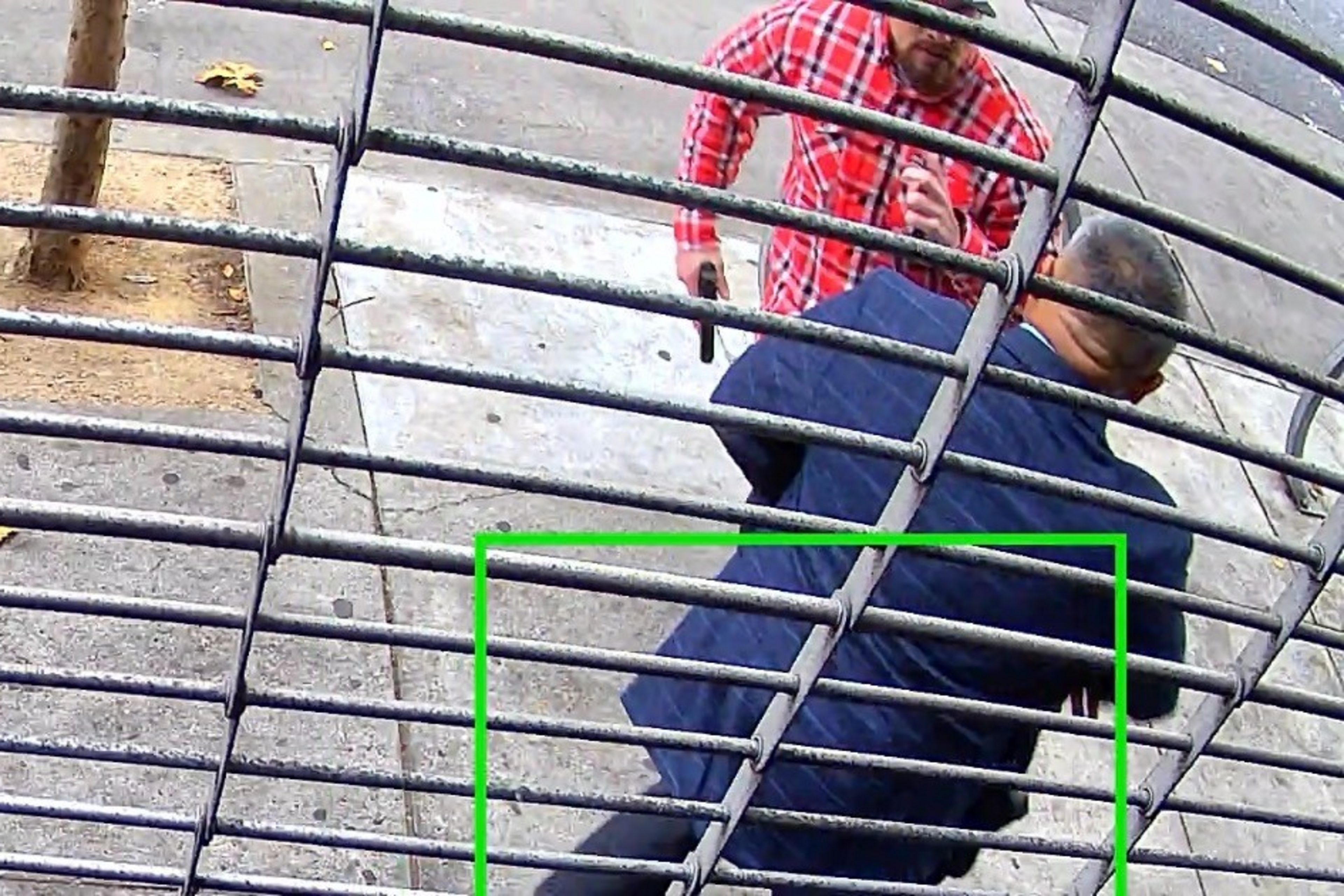 A man in a red plaid shirt points a handgun at another man in a blue jacket near a sidewalk and tree, viewed through metal bars.