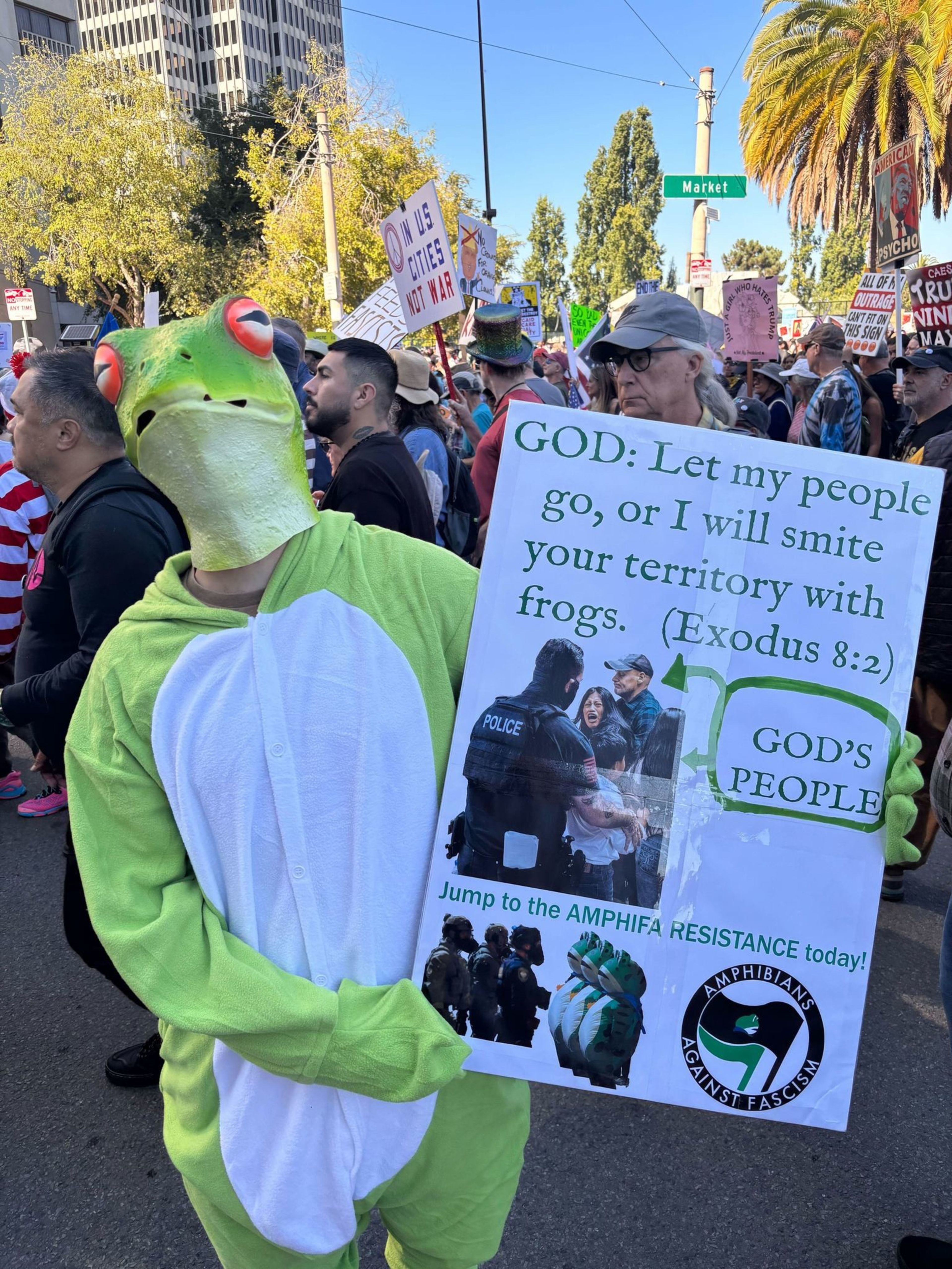 A person in a frog costume holds a sign quoting Exodus 8:2 and promoting “Amphibians Against Fascism” during a crowded protest at Market Street.