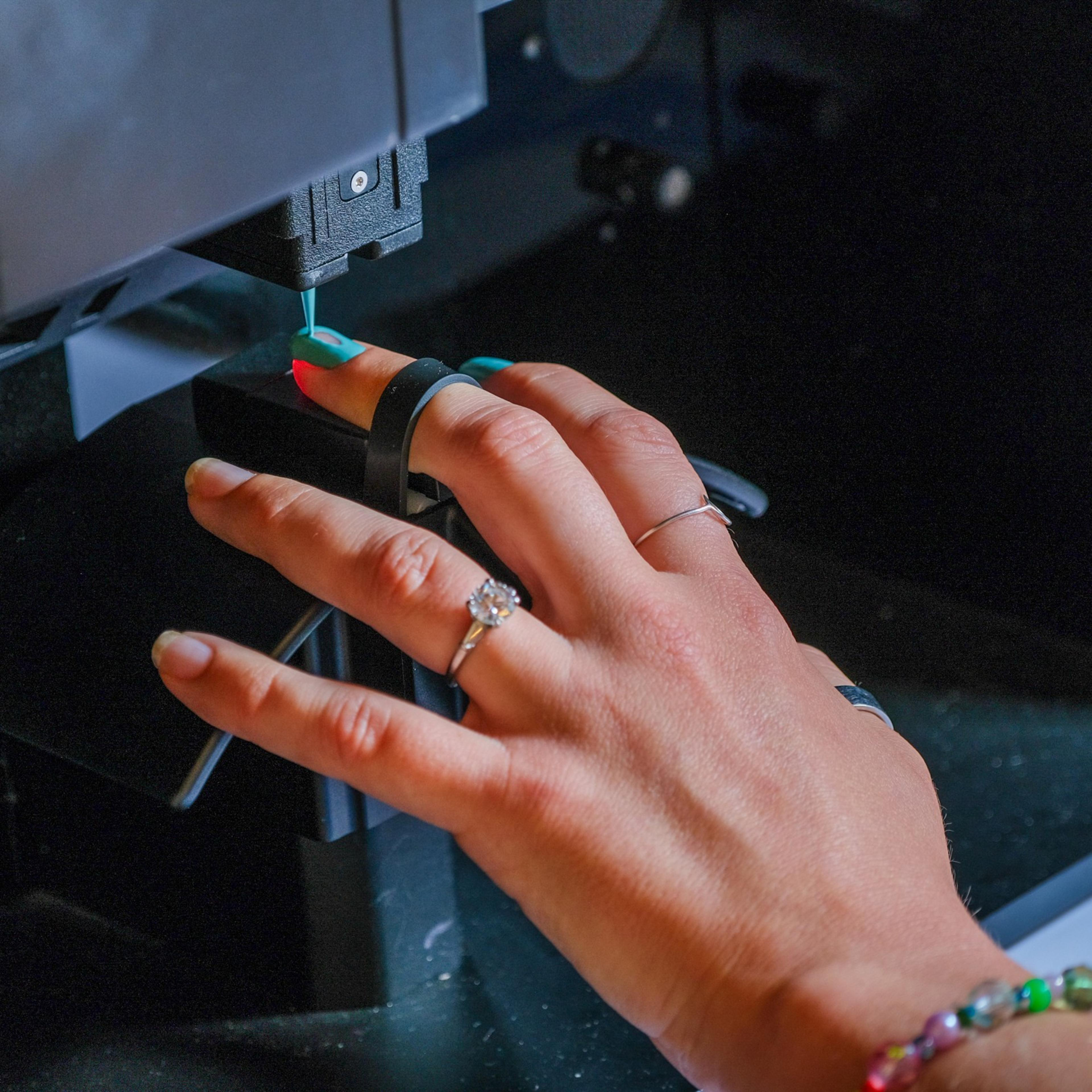 A hand with rings and bracelets is positioned under a small black machine, with a blue light or laser pointing at the nails, possibly for a nail treatment.