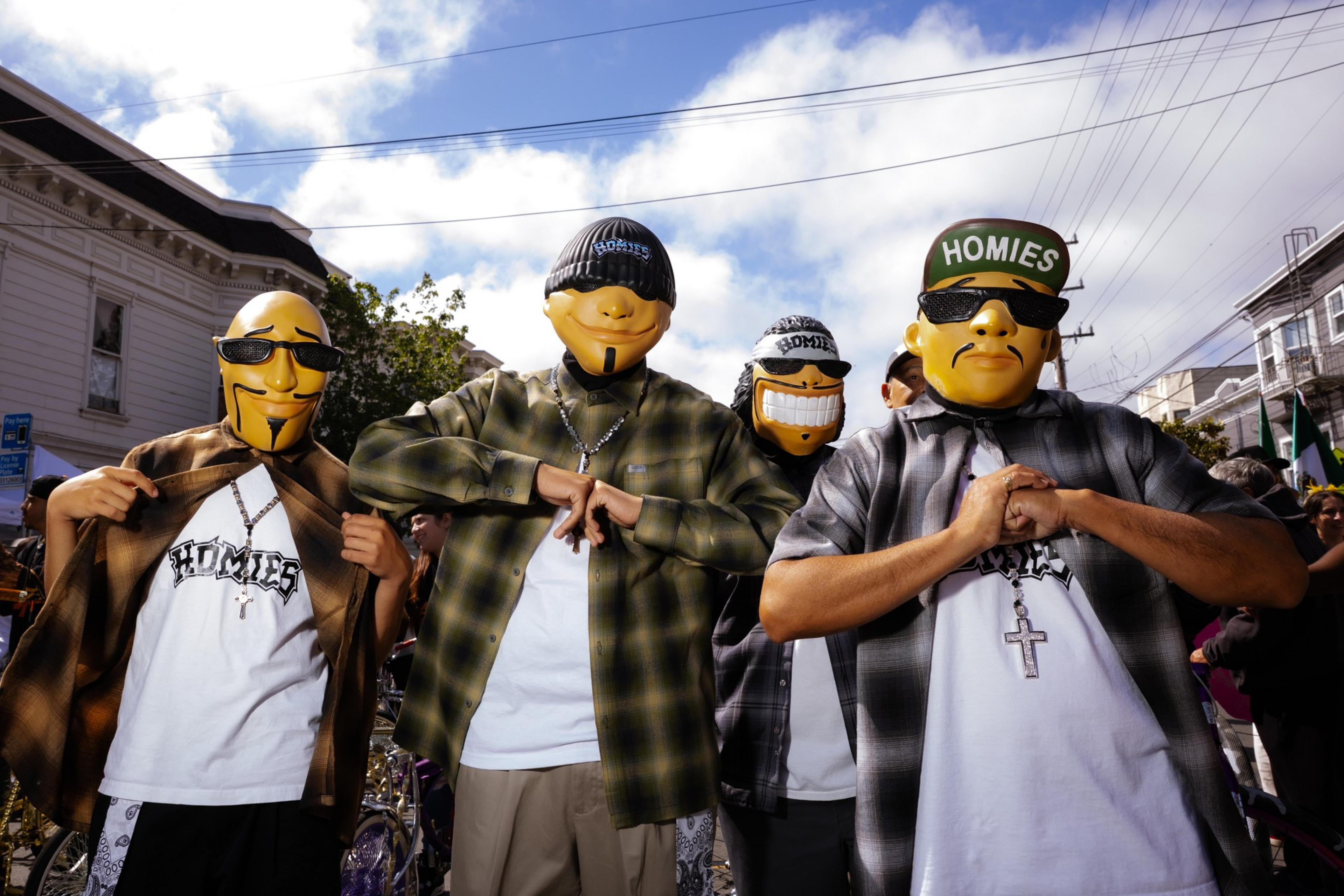 Four people wear cartoon-style masks and &quot;Homies&quot; hats or shirts. They stand outdoors, posing with hands clasped. It's a sunny day with a mix of clouds.