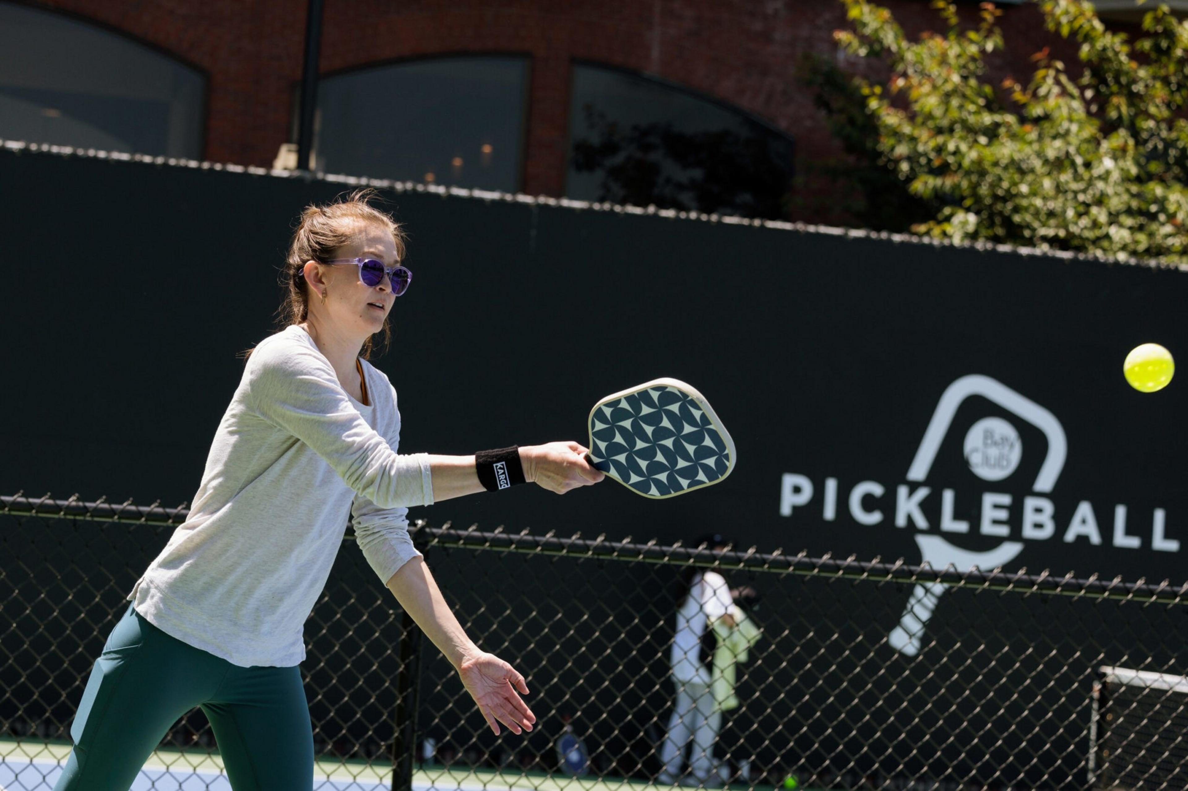A woman wearing sunglasses, a white long-sleeve shirt, and teal pants is hitting a yellow pickleball with a patterned paddle on an outdoor court.