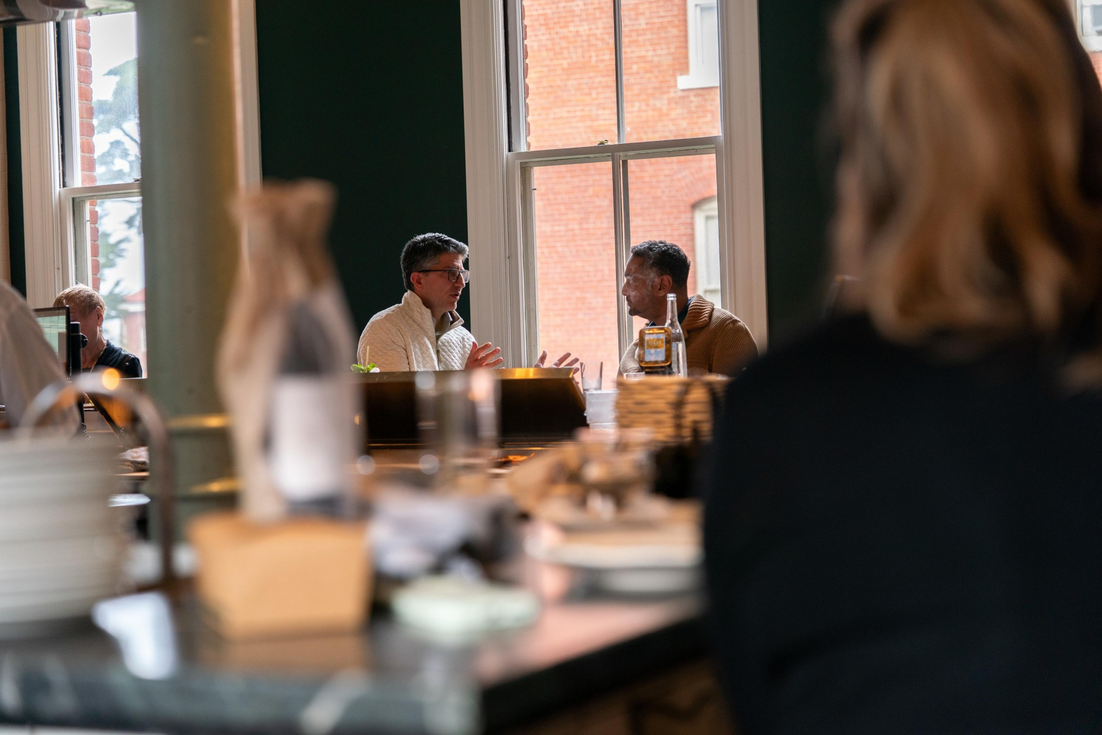 Two people sit at a table near a window, engaged in conversation. A clear bottle is on the table, with soft lighting in the background.
