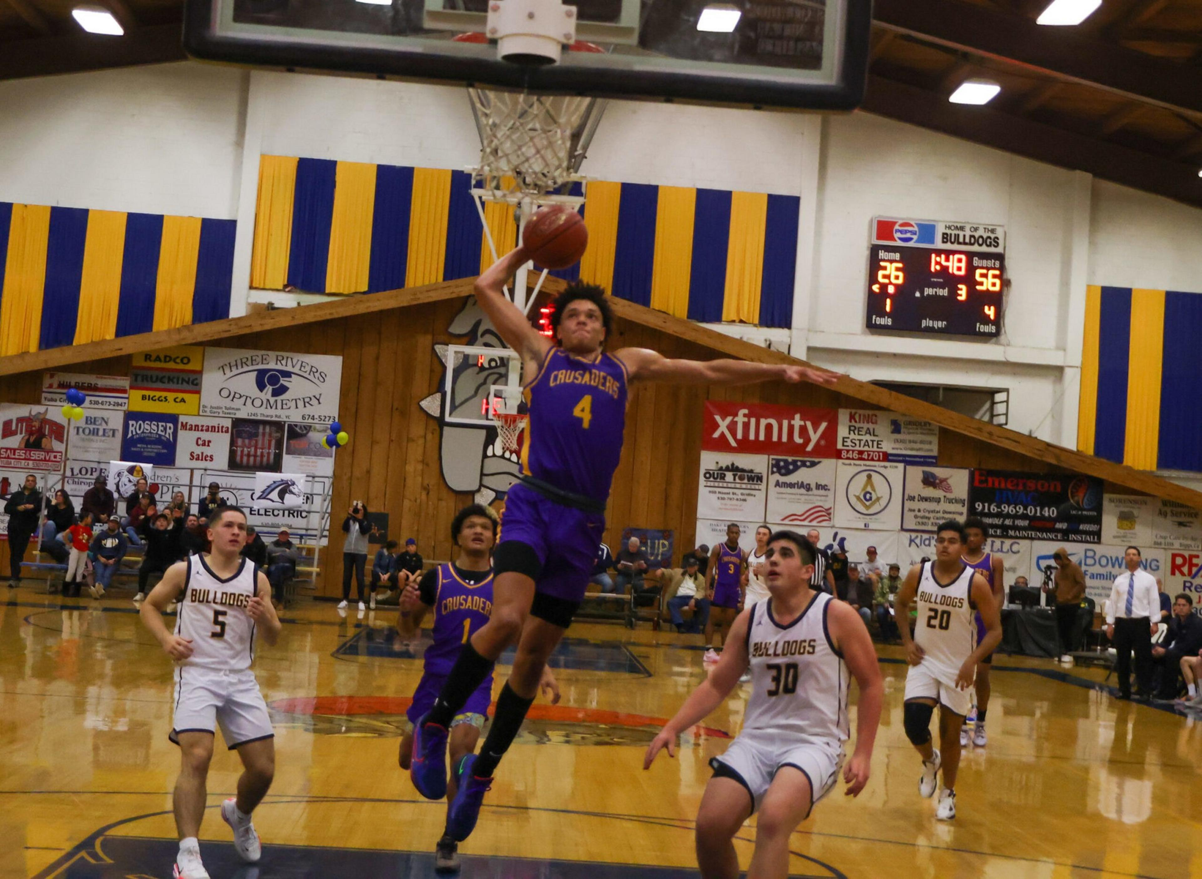 A basketball player in a purple "Crusaders" jersey leaps for a dunk while defenders in white "Bulldogs" jerseys watch on a gym court.