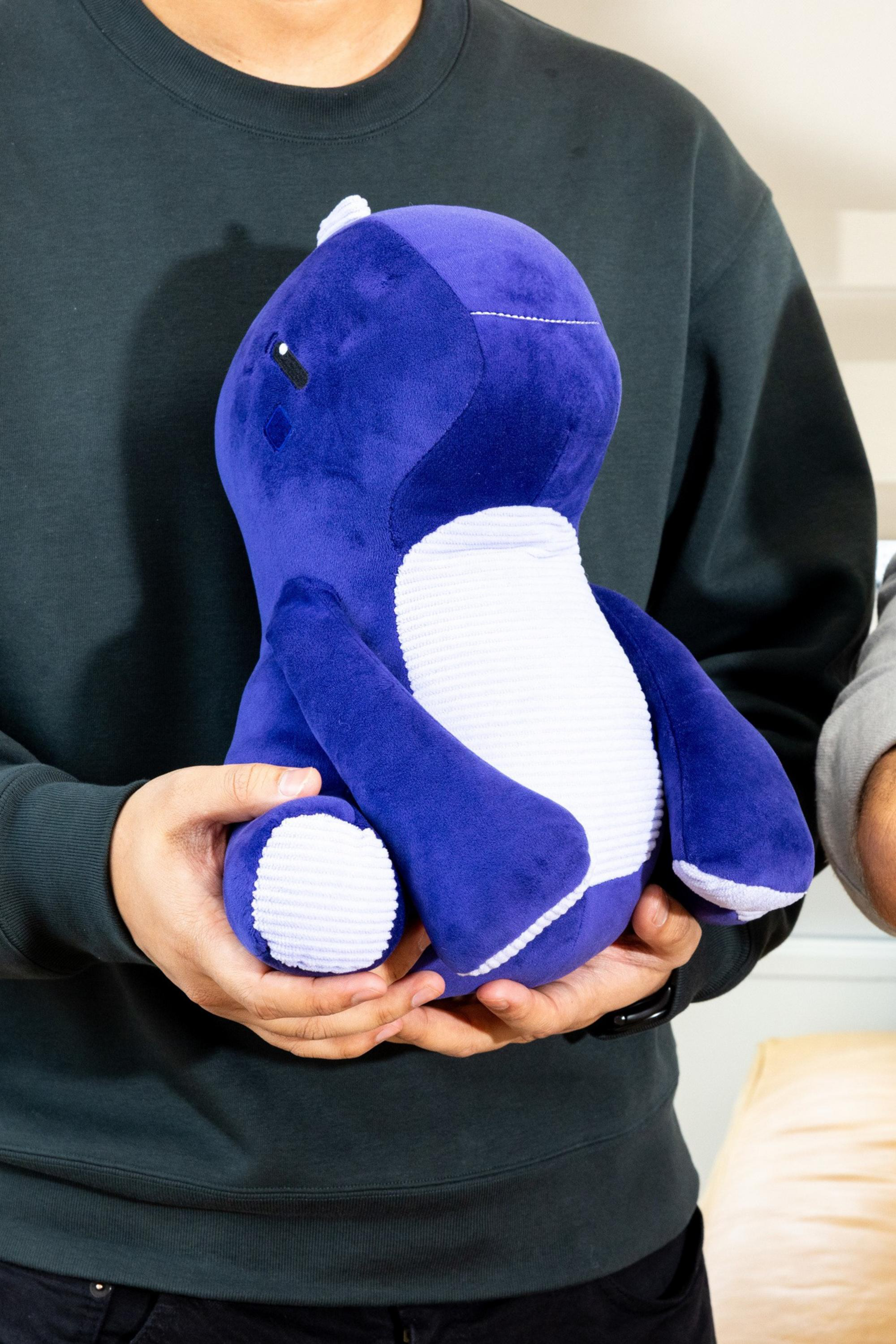 A person in a dark sweatshirt holds a plush toy resembling a blue and white dinosaur or dragon.