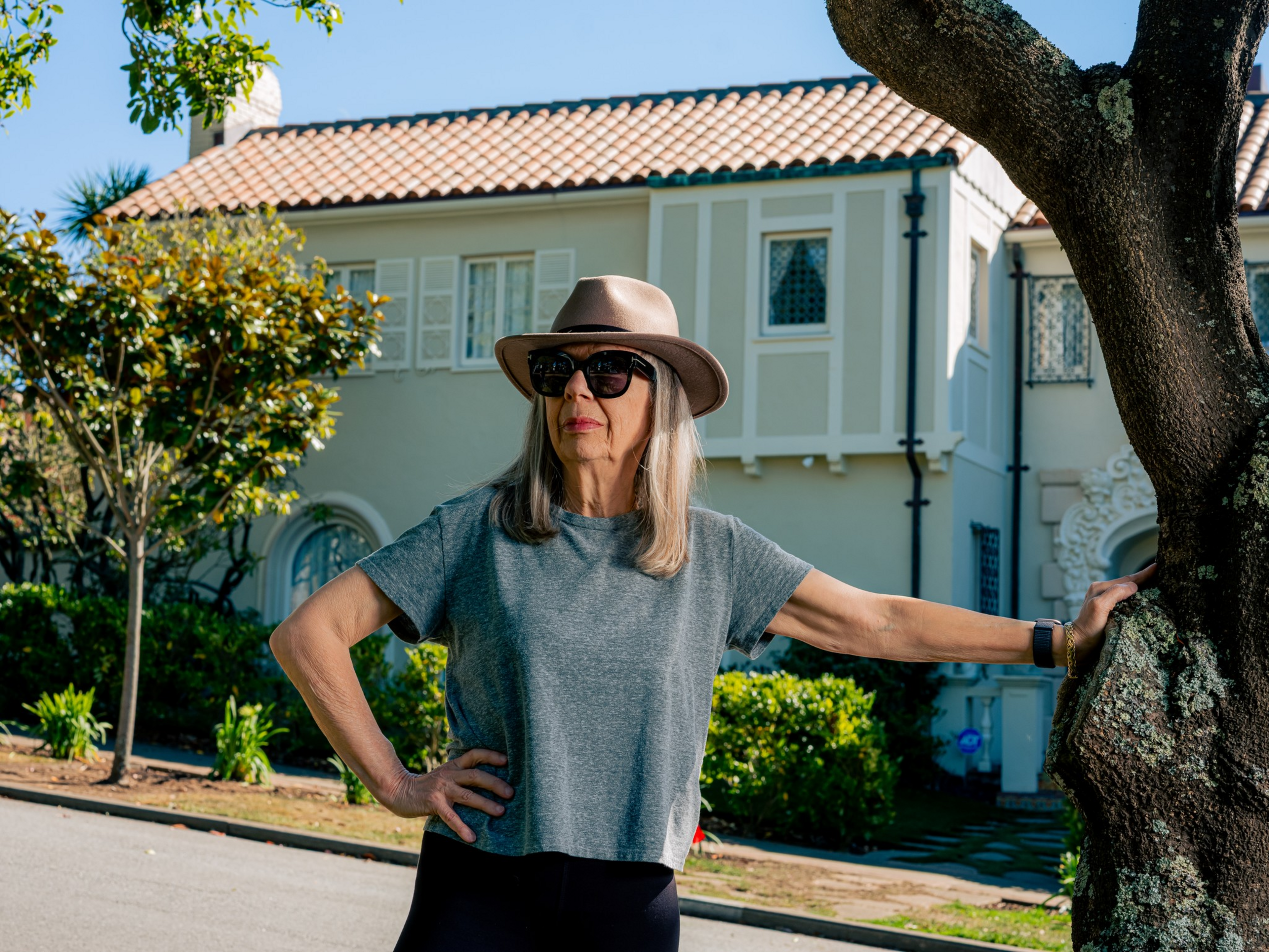 A woman wearing a gray t-shirt, sunglasses, and a beige hat stands by a tree, with one hand on her hip and a house with a tiled roof in the background.