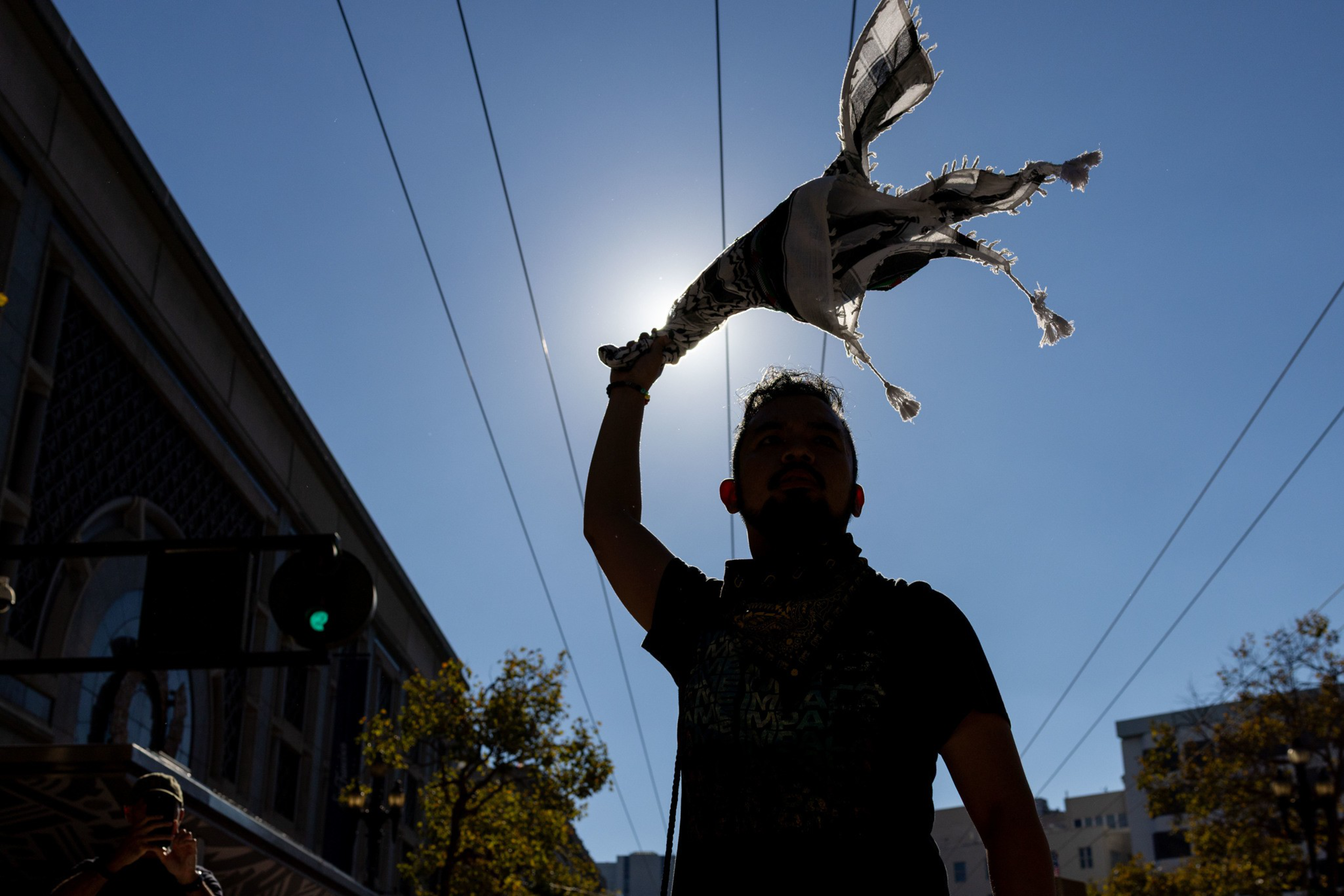 A person holds a frayed black-and-white scarf high above their head against a bright blue sky with sun backlighting them.