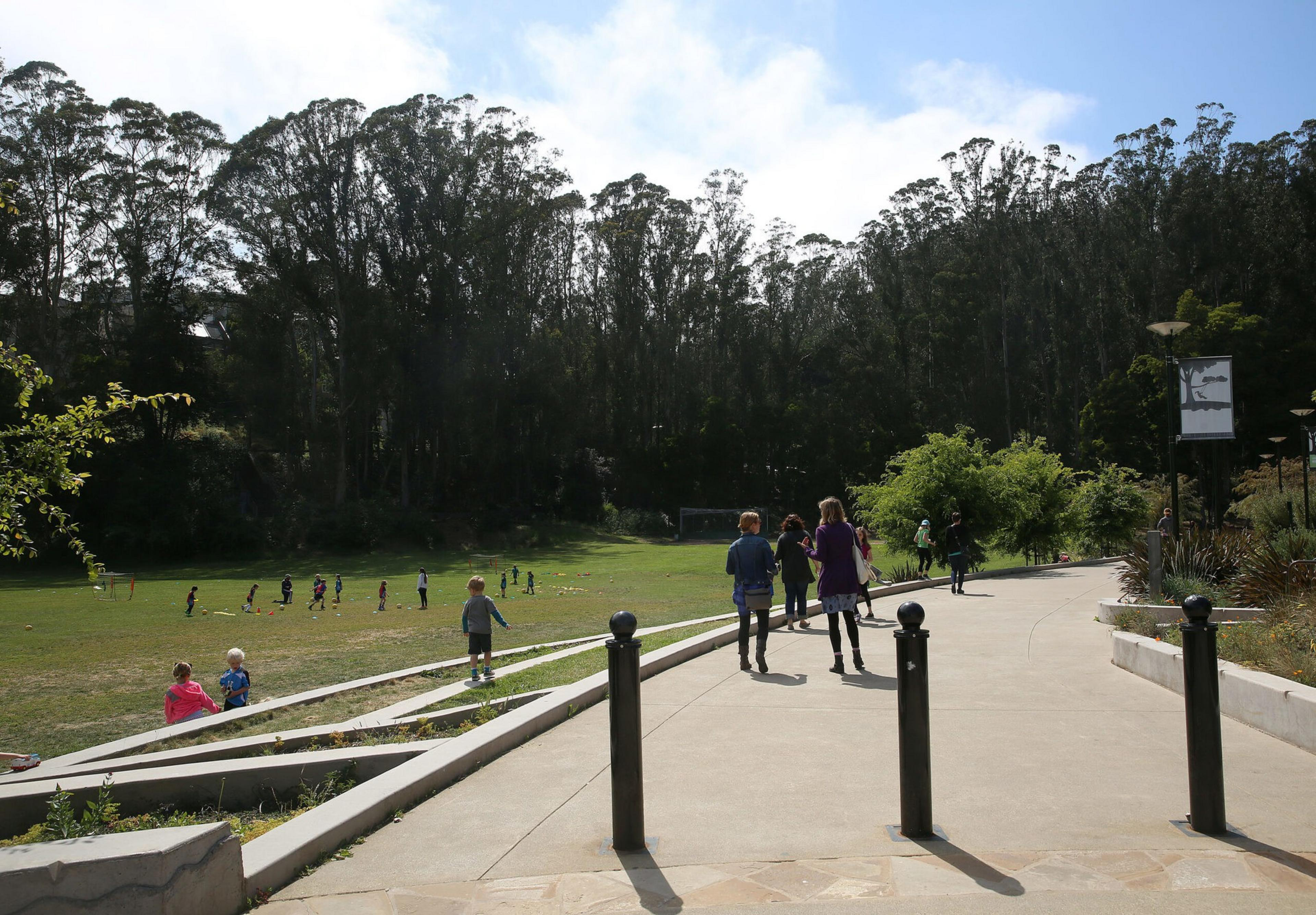 People walk along a paved path next to a grassy field where children play, with tall trees and a partly cloudy sky in the background.