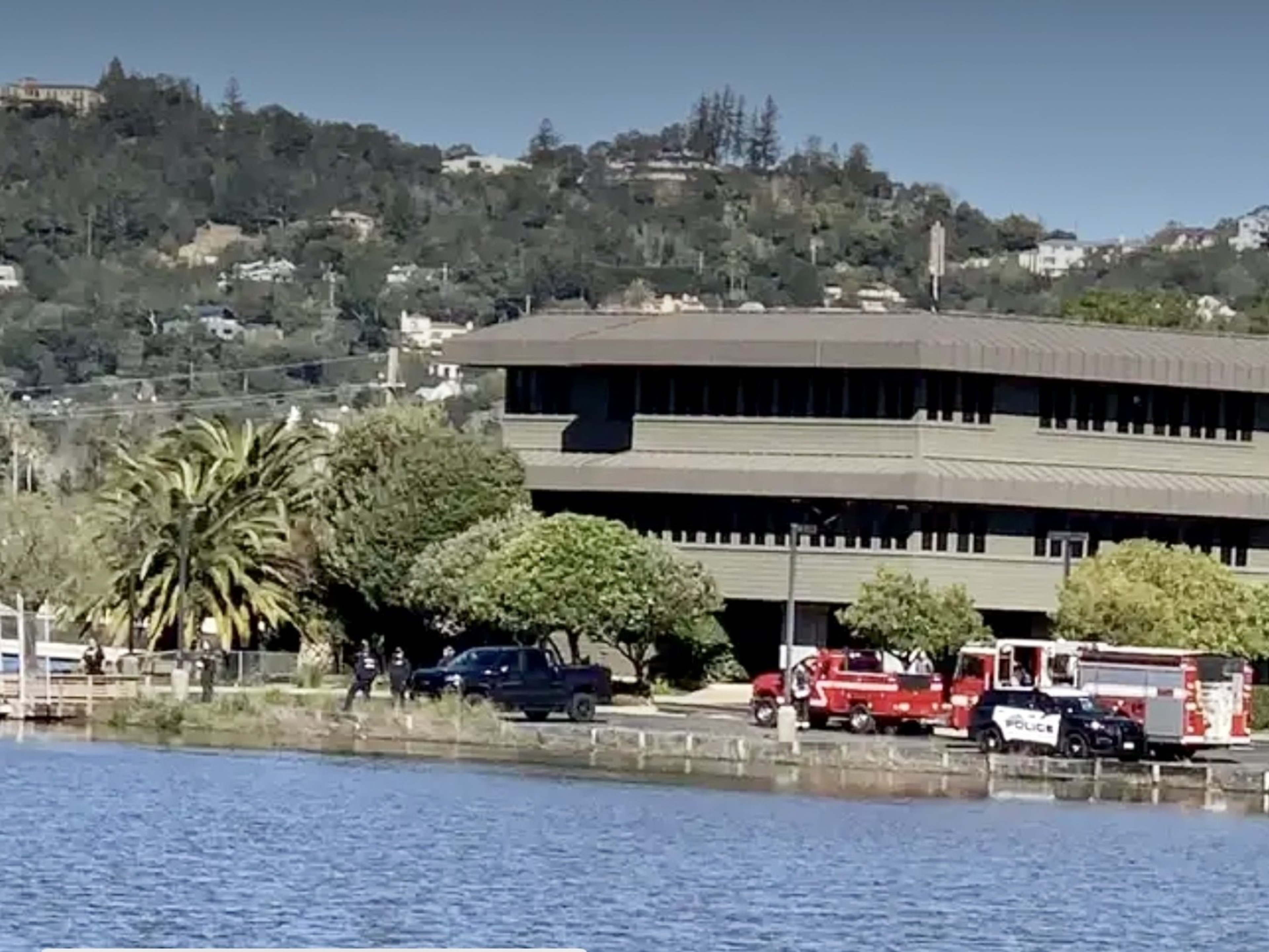 Fire trucks and a police vehicle by a building near water, with palm trees and hills in the background.