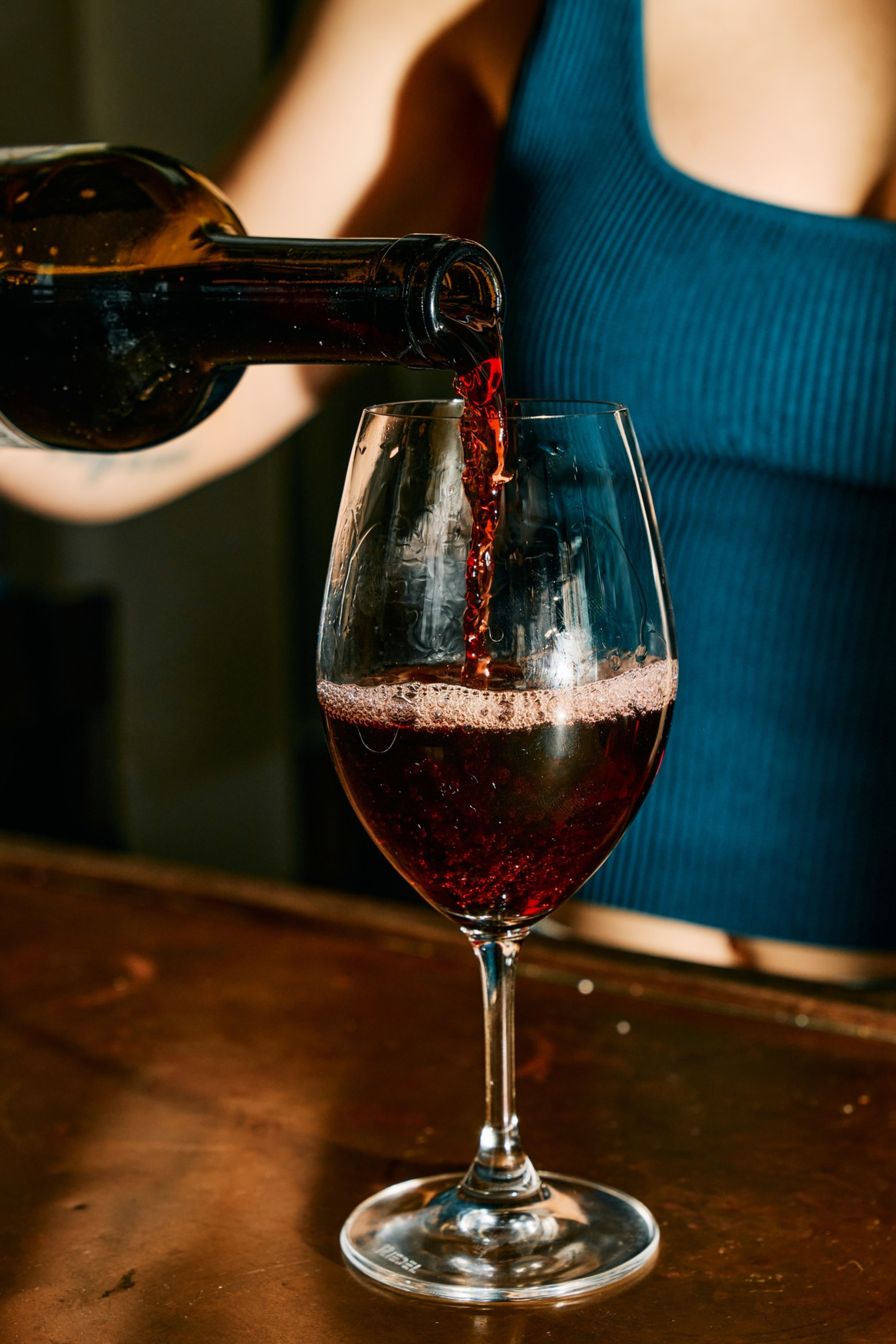 Red wine is being poured from a bottle into a glass held by a person wearing a blue sleeveless top, resting on a wooden surface.
