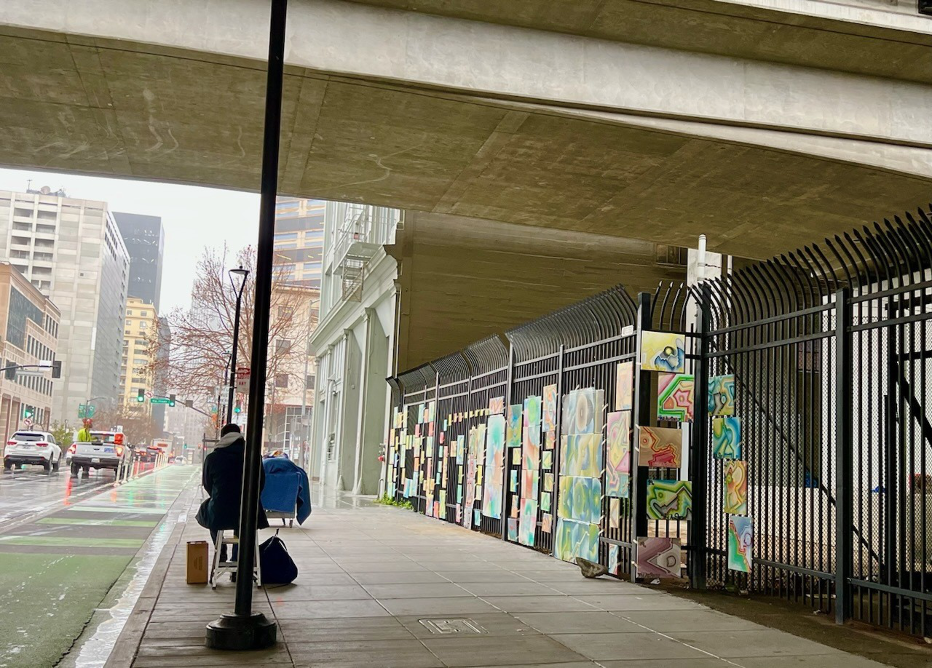A sidewalk under a concrete overpass features a black fence decorated with colorful abstract paintings, while two people sit near a lamppost on a rainy day.