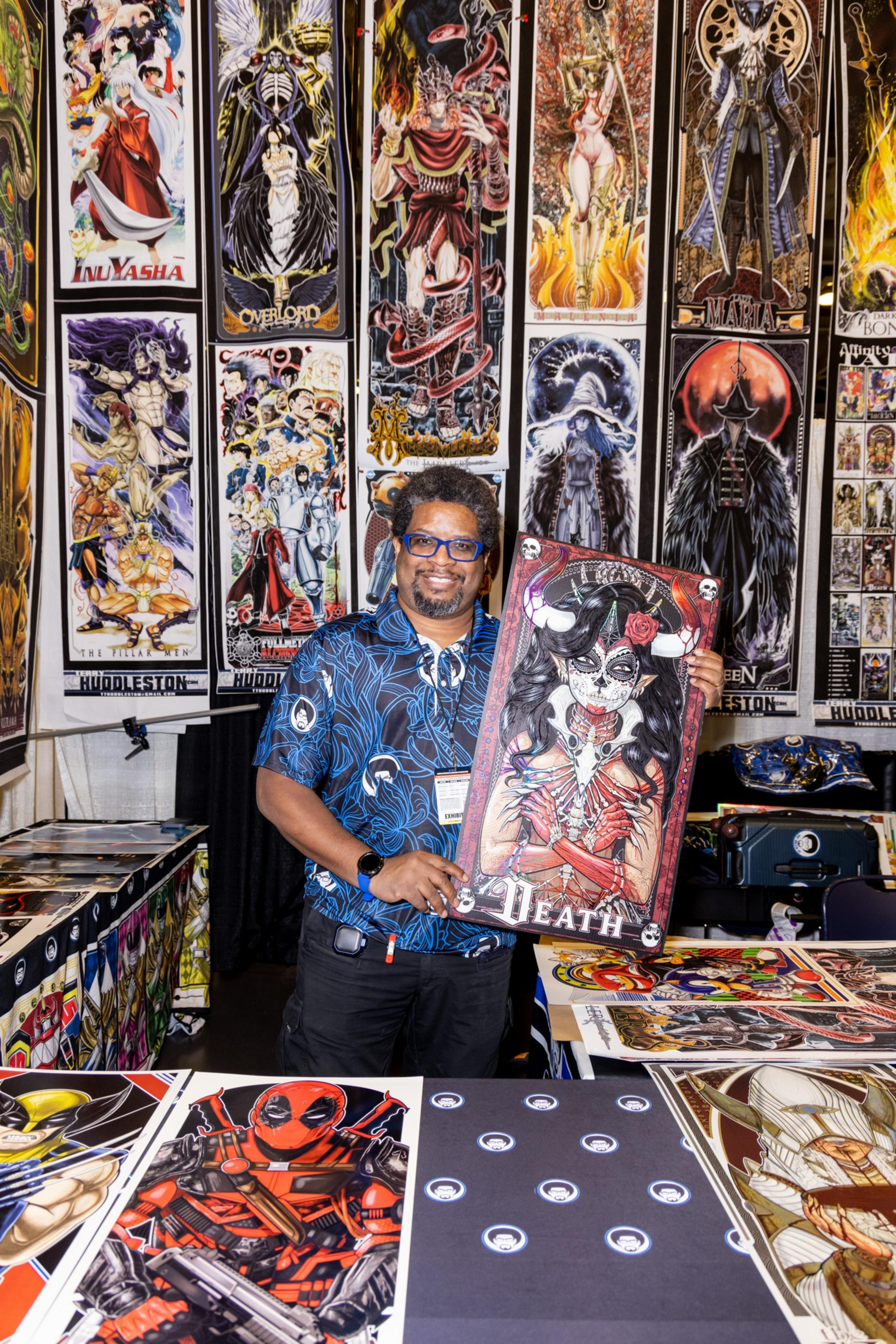 A person stands at a booth displaying vibrant anime and comic art. The individual holds a colorful, gothic-style poster titled &quot;DEATH,&quot; surrounded by more art pieces.