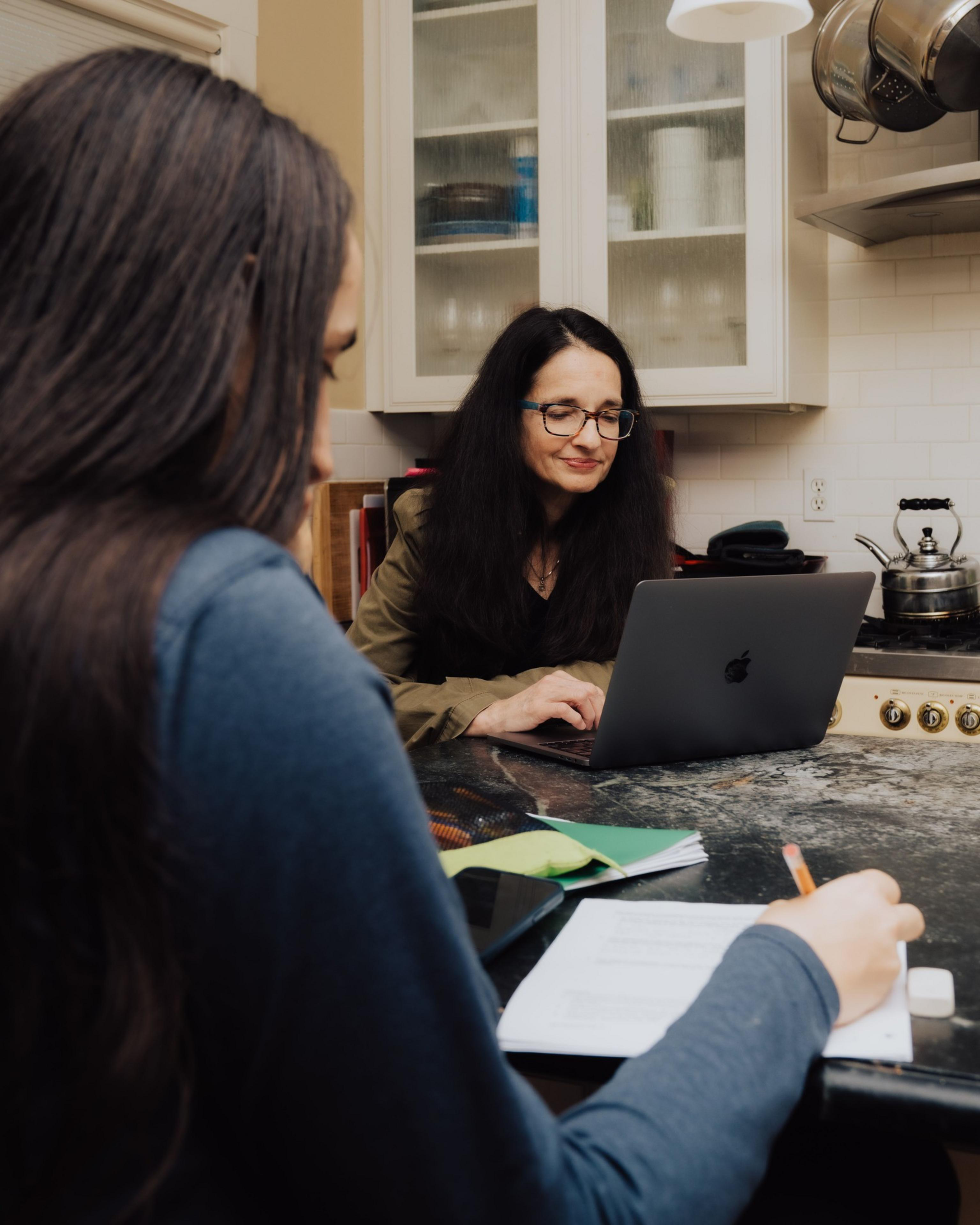 A woman with glasses uses a laptop in a kitchen. Another person is writing on paper at the table, surrounded by notebooks and a smartphone.
