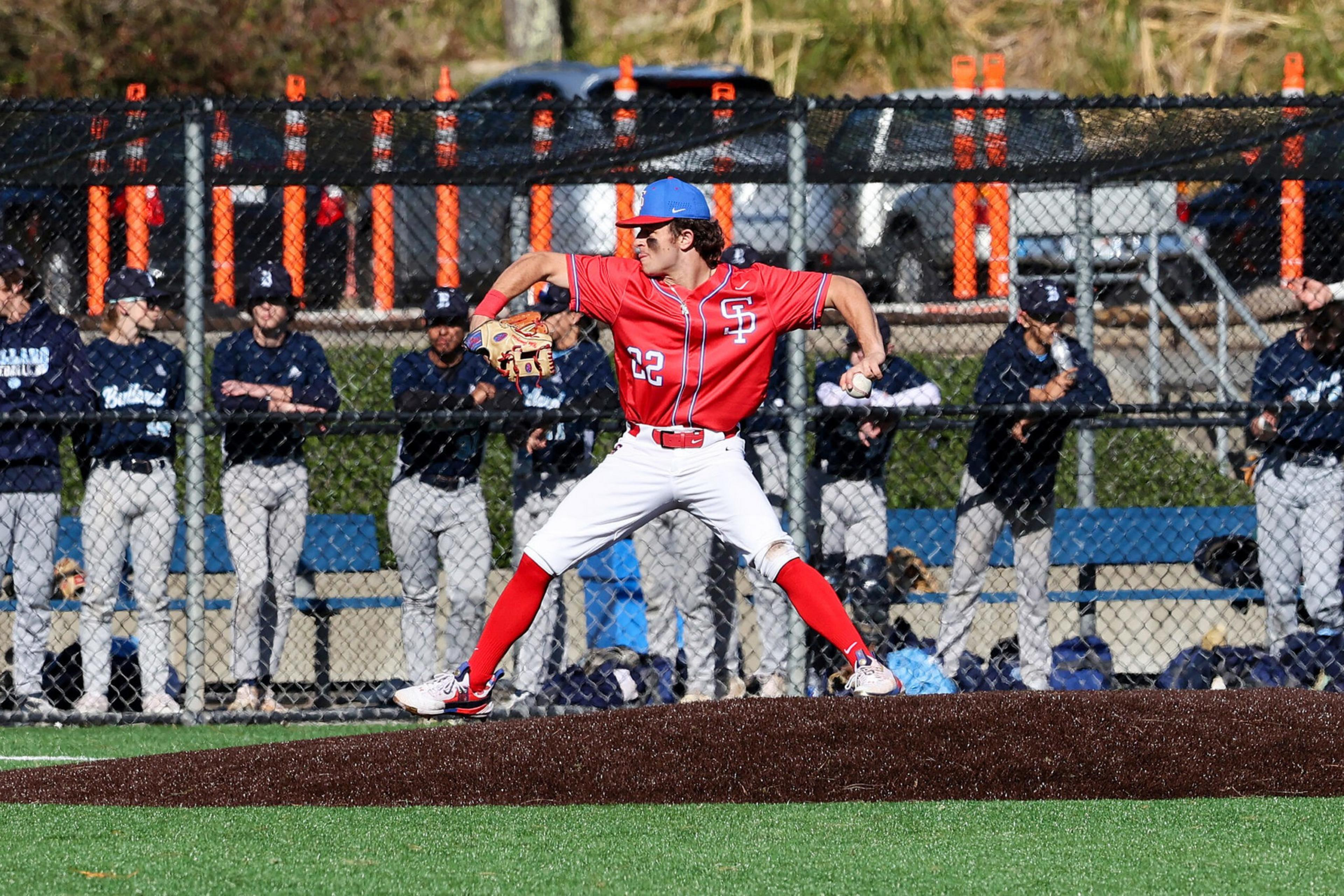 A baseball pitcher in a red uniform with number 22 winds up on the mound while players in navy uniforms watch from behind a fence.