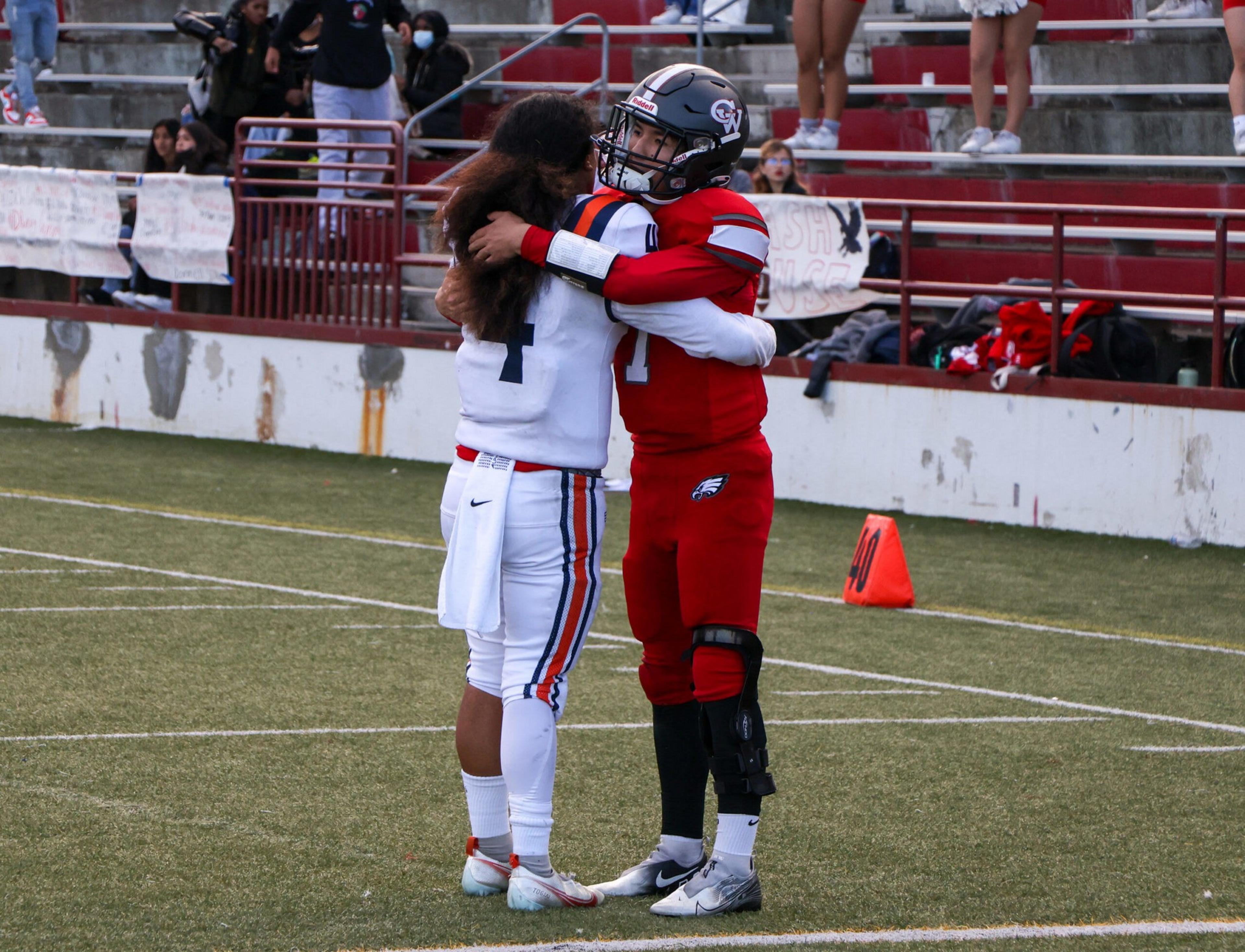 Two football players from opposing teams embrace on the field, with spectators and cheerleaders visible in the background.