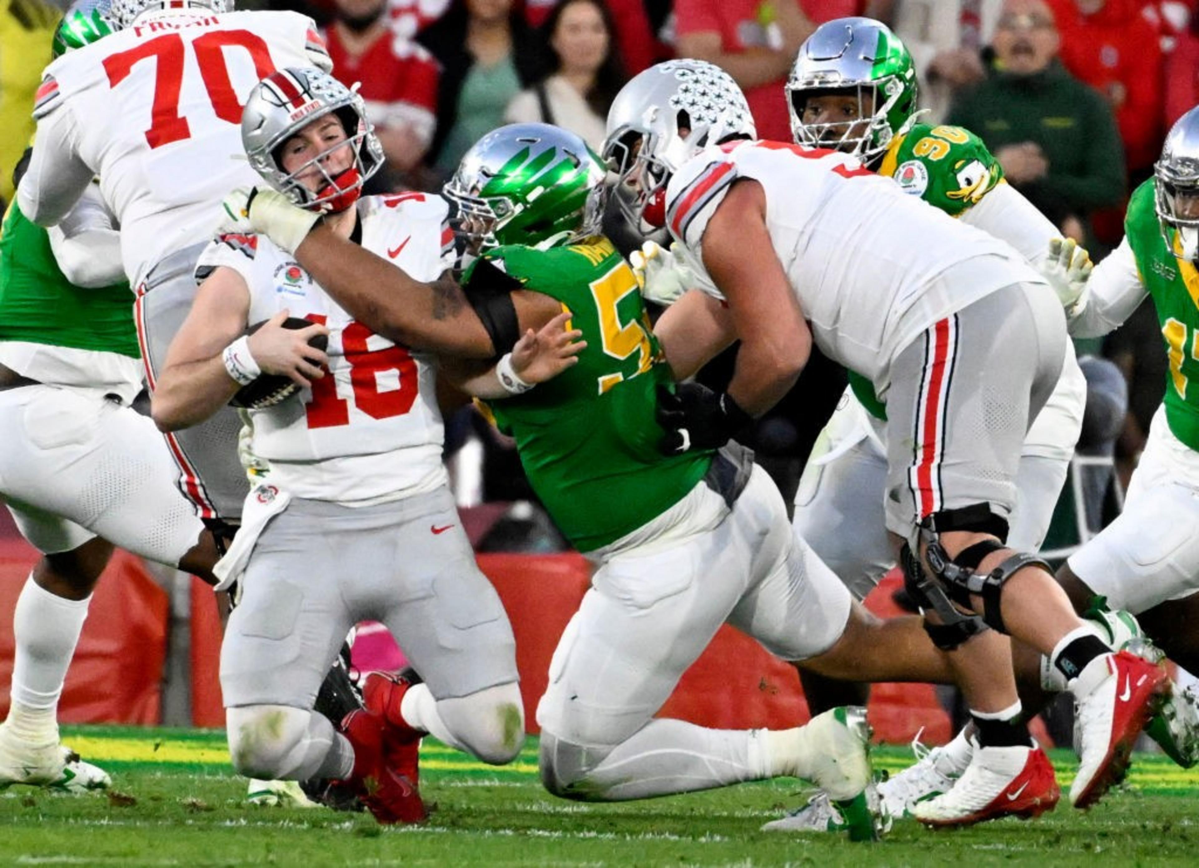 Two football teams wearing green and white uniforms are in action on the field. A player in white with the ball is being tackled by players in green.